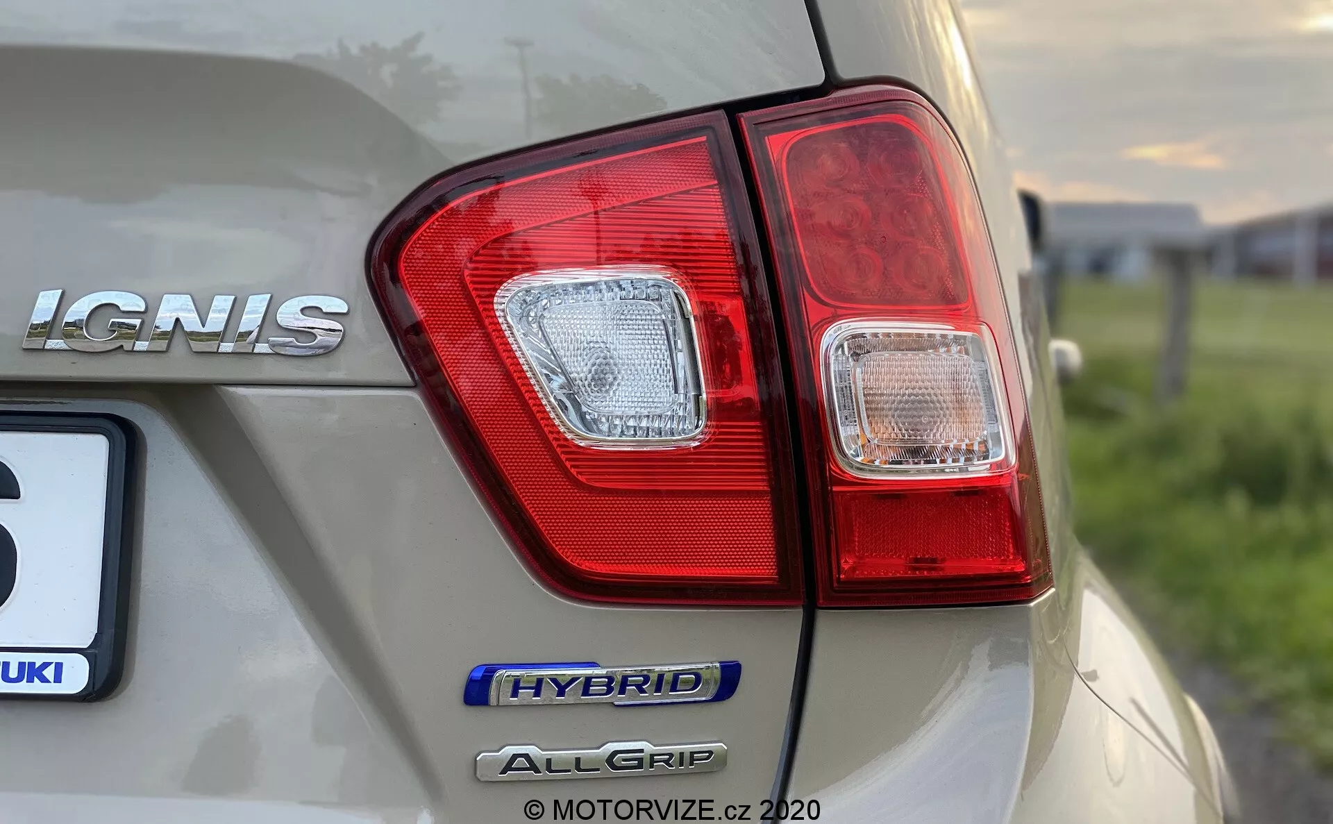 Bližnji, podroben posnetek zadnjega kota Suzuki Ignis 2020, osredotočen na zadnji svetlobni sklop in označbe modela iz nizke, rahlo kotne perspektive. Na desni strani avtomobila so jasno vidne oznake 'IGNIS', 'HYBRID' in 'ALLGRIP', kar poudarja hibridno tehnologijo in sistem štirikolesnega pogona. Avto svetle šampanjske barve ima sijoč zaključek z odsevi okolja, slika pa zajema ostre podrobnosti zadnjega svetlobnega sklopa z rdečimi in prozornimi segmenti, pri čemer ohranja majhno globinsko ostrino.