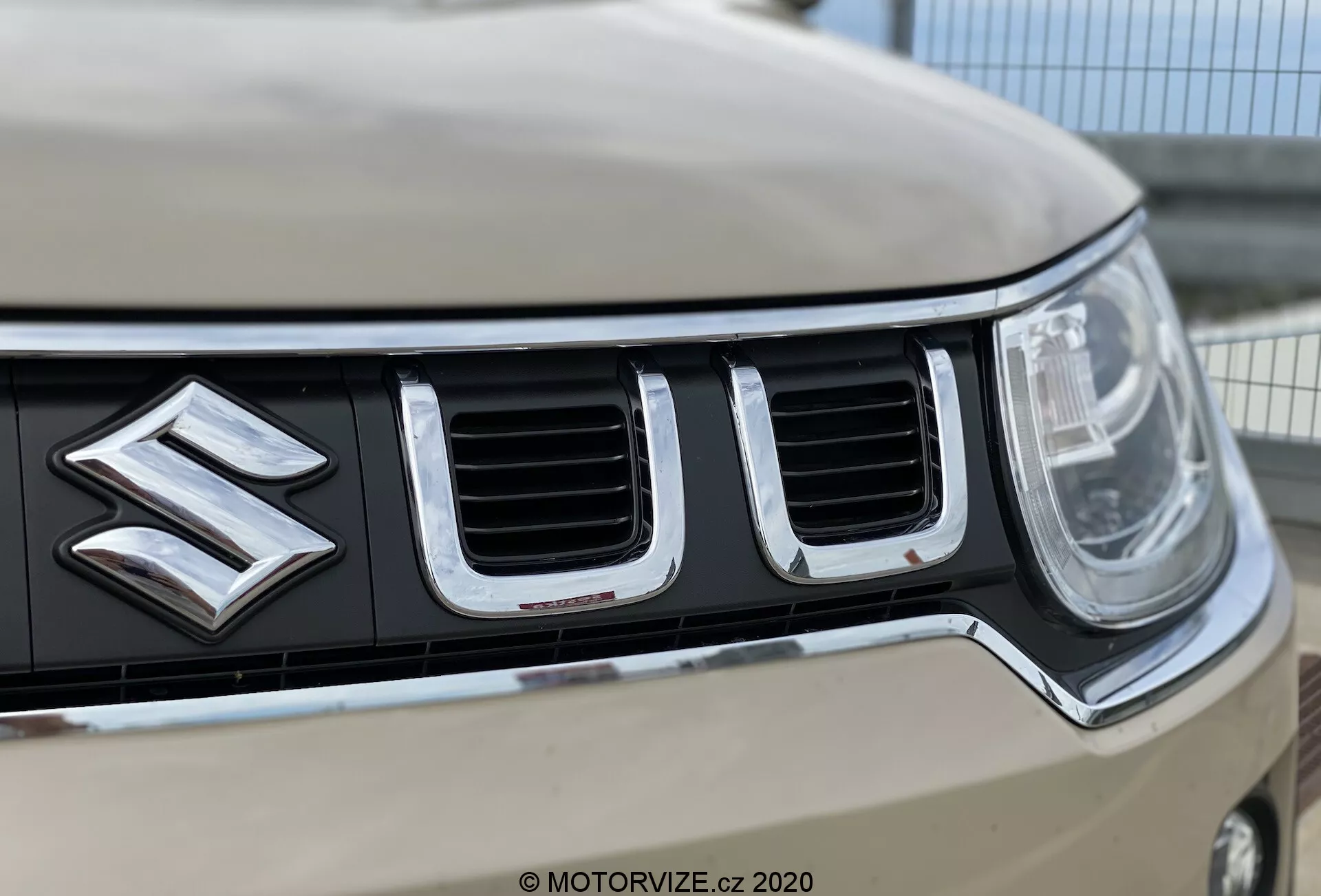 Od blizu posnetek sprednje rešetke in emblema avtomobila Suzuki Ignis 2020, poudarja izrazit logotip Suzuki, navpično usmerjene kromirane reže, levo žaromet s sodobno LED ali HID tehnologijo in svetlo karoserijo. Plitva globinska ostrina poudarja podrobnosti rešetke in logotipa, medtem ko zabriše oddaljene robove in ozadje.