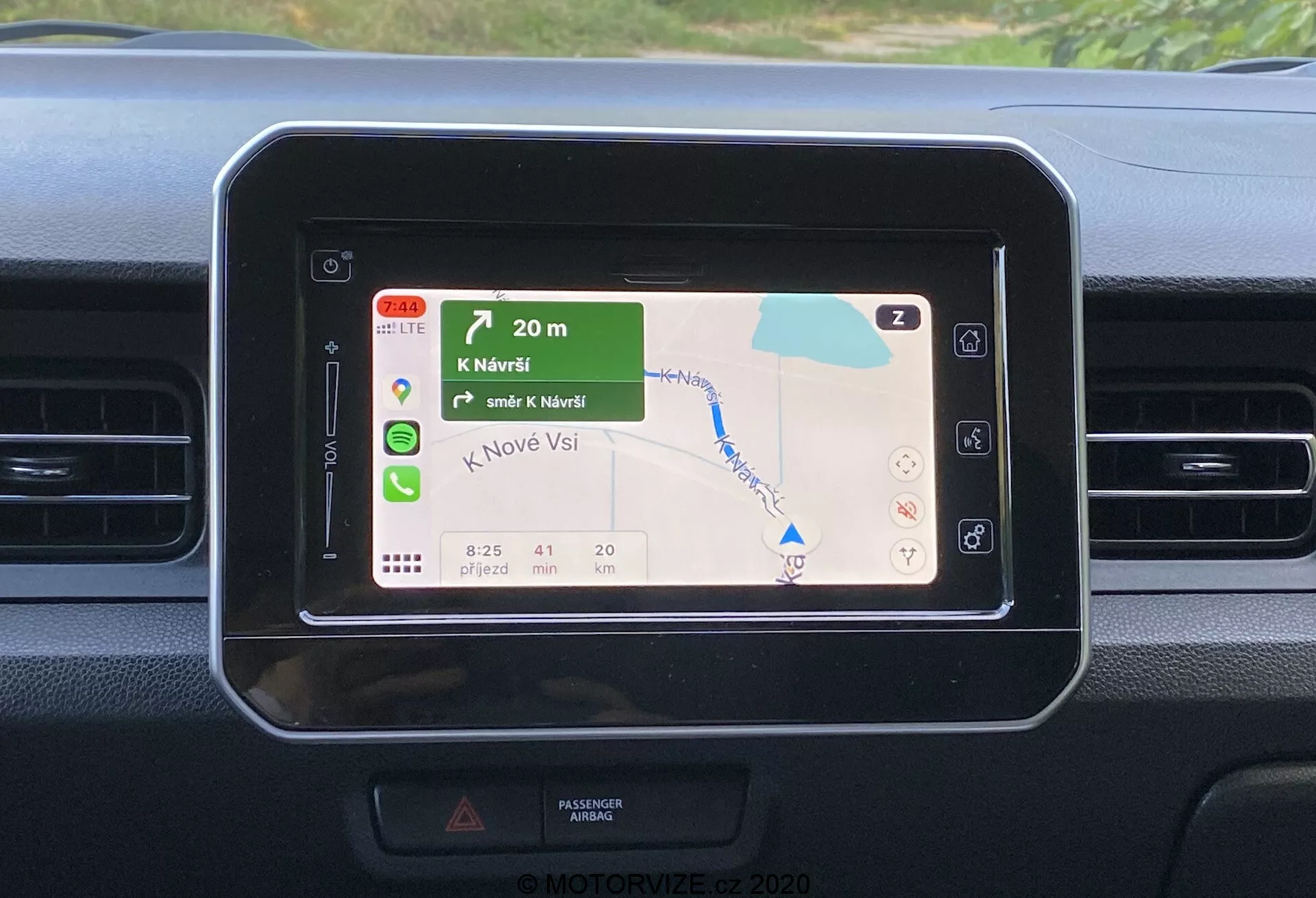 Pogled od blizu na sredinsko armaturno ploščo Suzuki Ignis 2020, osredotočen na pravokotno zaslon infozabavnega sistema z zaobljenimi vogali, ki prikazuje barvit GPS navigacijski zemljevid z zelenimi pokrajinami in modrimi vodnimi telesi, ki kaže navodila za zavijanje, ETA, razdaljo in omejitve hitrosti. Armaturna plošča ima preproste oblikovne elemente z vodoravnimi zračniki v bližini, nasprotujočimi se teksturami materialov in nakazuje upravljanje na dotik ali daljinsko upravljanje brez vidnega zunanjega konteksta.