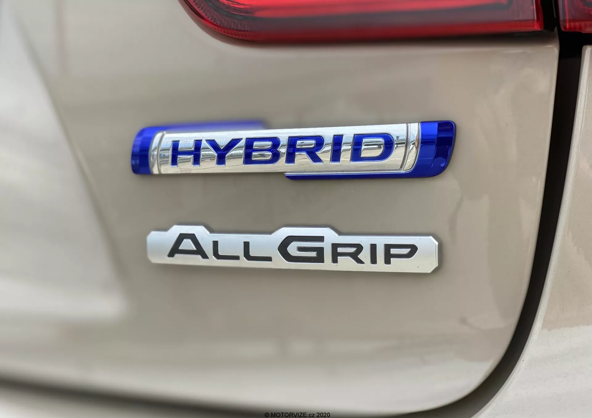 Bližnji posnetek zadnjih značk Suzuki Ignis 2020, ki poudarja lastnosti 'HYBRID' in 'ALLGRIP'; jasen, neposreden posnetek z majhno globinsko ostrino, ki poudarja okolju prijazno hibridno tehnologijo in napreden sistem pogona na vsa kolesa; naravna osvetlitev z ostrimi podrobnostmi in rahlimi odsevi.