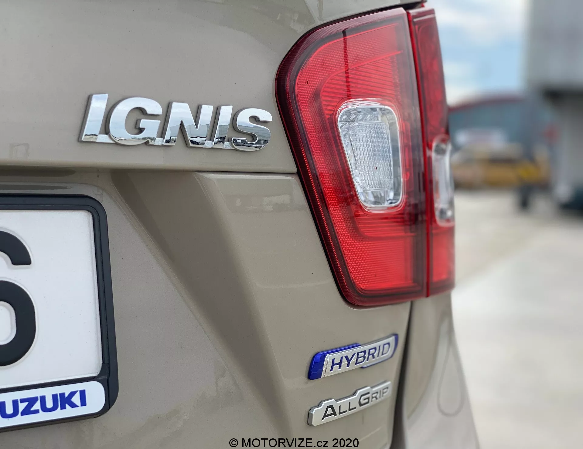 Podrobni posnetek zadnjega dela vozila Suzuki Ignis 1.2 Dualjet Hybrid Club 2020, ki poudarja zadnjo desno luč z rdečimi in prozornimi deli, kromirano oznako modela 'IGNIS', značko 'HYBRID' in značko 'ALLGRIP', ki nakazuje hibridno tehnologijo in sistem štirikolesnega pogona. Znak Suzuki je delno viden tudi nad registrsko tablico, avto pa je v bež ali svetlo rjavi kovinski barvi. Ozadje je zamegljeno, da poudari blagovno znamko in tehnološke značilnosti vozila.