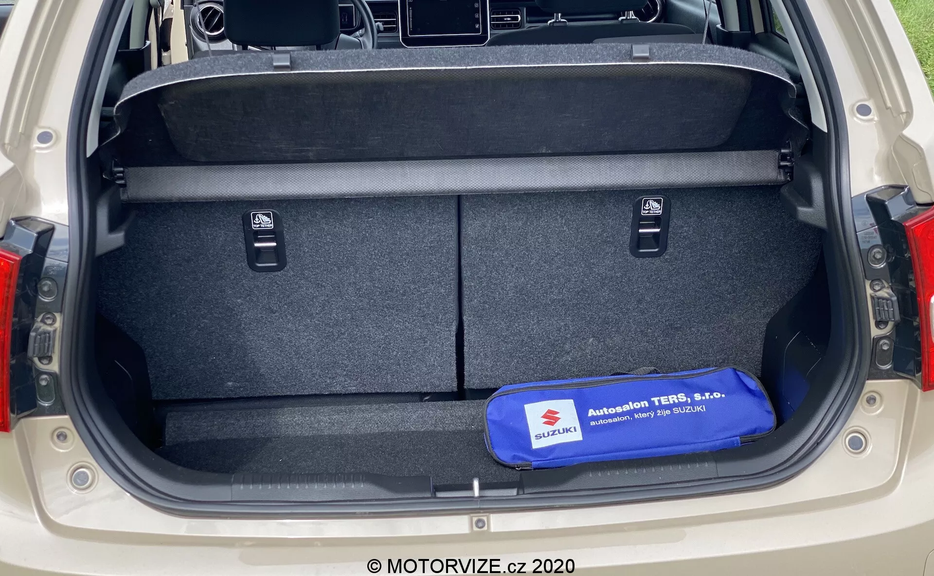 Prtljažnik avtomobila Suzuki Ignis 2020, gledan s hrbtne perspektive, prikazuje veliko pravokotno shranjevalno območje s preprogo na tleh in straneh, majhne vdolbine za predmete, izvlečki za zlaganje zadnjih sedežev, modri varnostni ali orodjarni komplet na tleh, viden moderen dizajn zadnjih luči ob straneh prtljažnika.