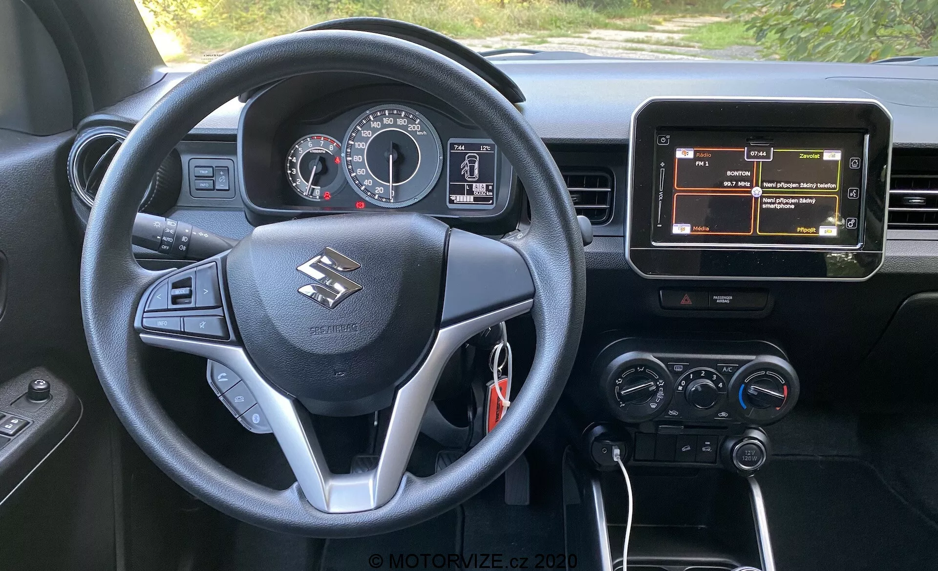 Pogled z voznikovega sedeža v notranjost Suzuki Ignis 1.2 Dualjet Hybrid Club 2020, ki prikazuje večfunkcijski volan s krmilniki za avdio, telefon in informacije o vozilu, instrumentno ploščo z analognimi merilniki in osrednjim digitalnim zaslonom, osrednji zaslon infozabave z navigacijo in gumbe za upravljanje klimatske naprave. Vozilo ima samodejni menjalnik, kar je nakazano s črko 'P' na prestavni ročici, kar nakazuje, da je parkirano, in črno notranjost s srebrnimi poudarki.