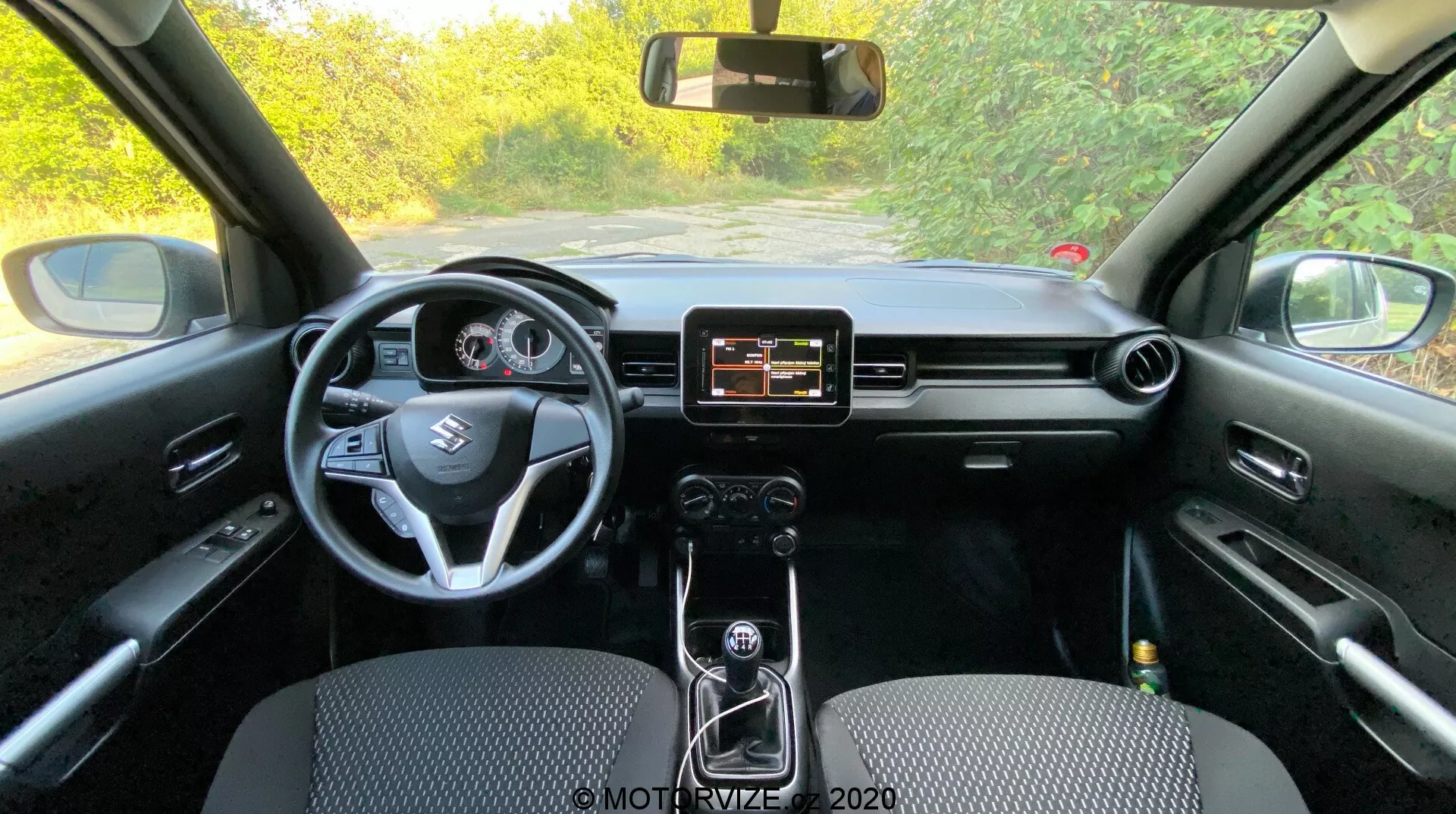 Notranji pogled na Suzuki Ignis 2020 iz zadnjega sedeža prikazuje armaturno ploščo, volan, informacijsko-razvedrilni sistem in instrumentno ploščo. Sredinska konzola z ročnim menjalnikom, črna notranjost z različnimi teksturami in materiali.