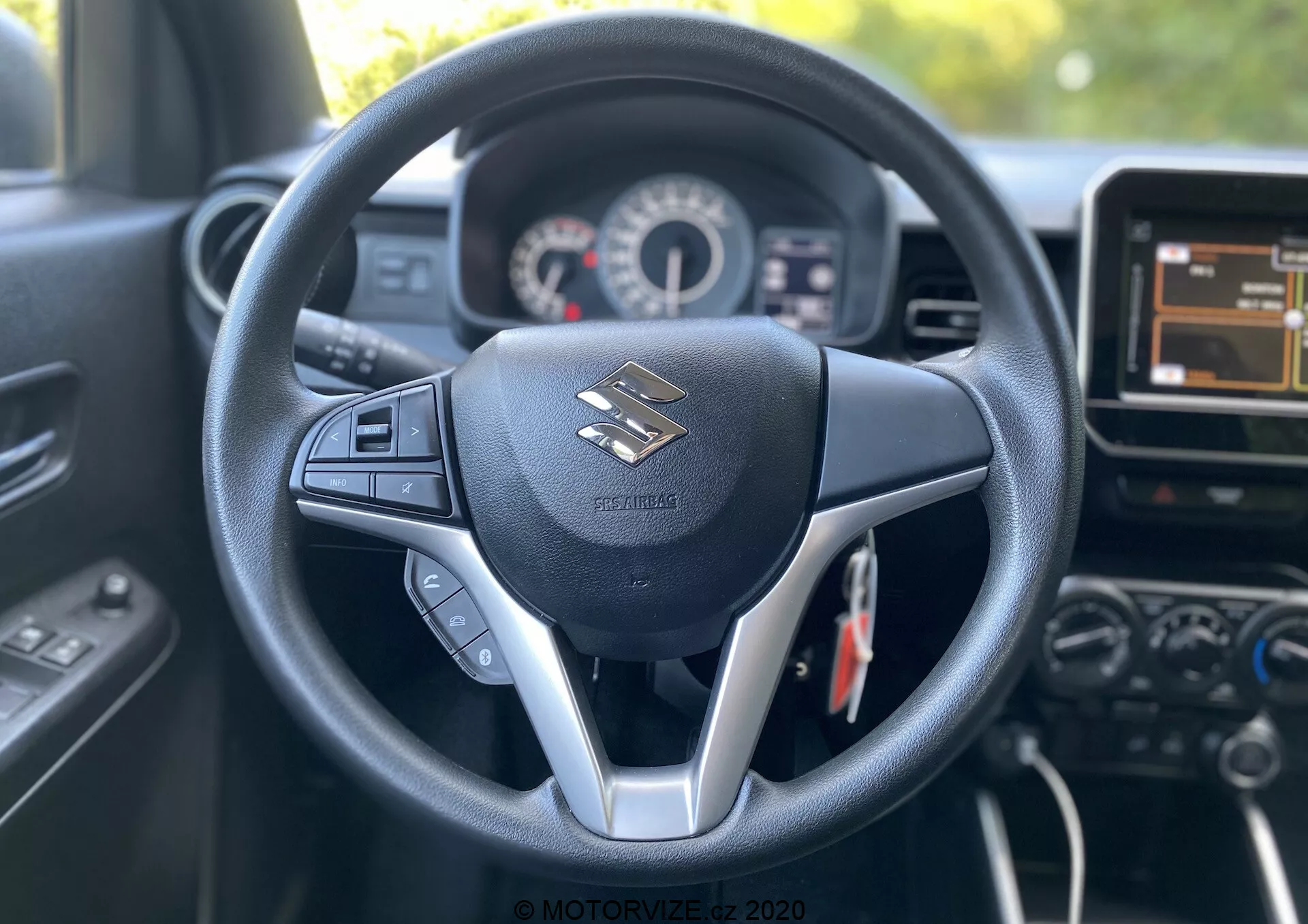 Pogled v notranjost modela Suzuki Ignis 2020 iz perspektive voznikovega sedeža, ki prikazuje osredotočeno volansko kolo s trokrakim dizajnom in srebrnimi detajli. Volan ima vgrajene kontrole za avdio in verjetno tempomat. Za njim je videti instrumentno ploščo s hitrostno in tahometrom ter majhnim digitalnim zaslonom. Slika zajema tudi del osrednje konzole, vključno z zaslonom na dotik infotelematskega sistema in dodatnimi kontrolami. Notranjost izraža sodoben dizajn s črno barvno shemo, sintetičnim usnjem in plastičnimi komponentami, ki poudarjajo udobje in tehnologijo za voznika.