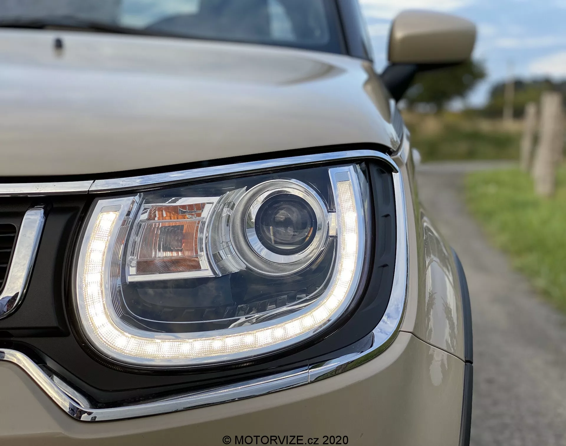 Podroben pogled na sprednjo levo enoto žarometa Suzuki Ignis 2020, z osrednje nameščeno glavno lučjo v slogu projektorja, LED dnevnimi voznimi lučmi, ki obdajajo ukrivljeno obliko žarometa, oranžnim smernikom na vrhu in svetlo obarvano karoserijo, verjetno srebrno ali belo, obdaja žaromet. Na levi strani je videti del kromirane maske, z majhno globino polja, ki poudarja zapleten dizajn in svetlobno tehnologijo.