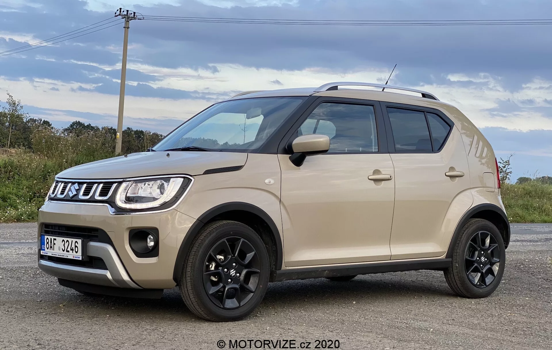Sprednji tridelni pogled na Suzuki Ignis 1.2 Dualjet Hybrid Club 2020, ki prikazuje njegovo sodobno kompaktno zasnovo SUV z dvobarvno karoserijo, izrazito rešetko in žarometi ter robustnimi kolesi. Sodobno estetiko vozila poudarjajo črna streha, ogledala, stranski okrasi in velike nizkoprofilne pnevmatike, kar vse kažejo na visoko oddaljenost od tal, primerno za različne vozne pogoje.