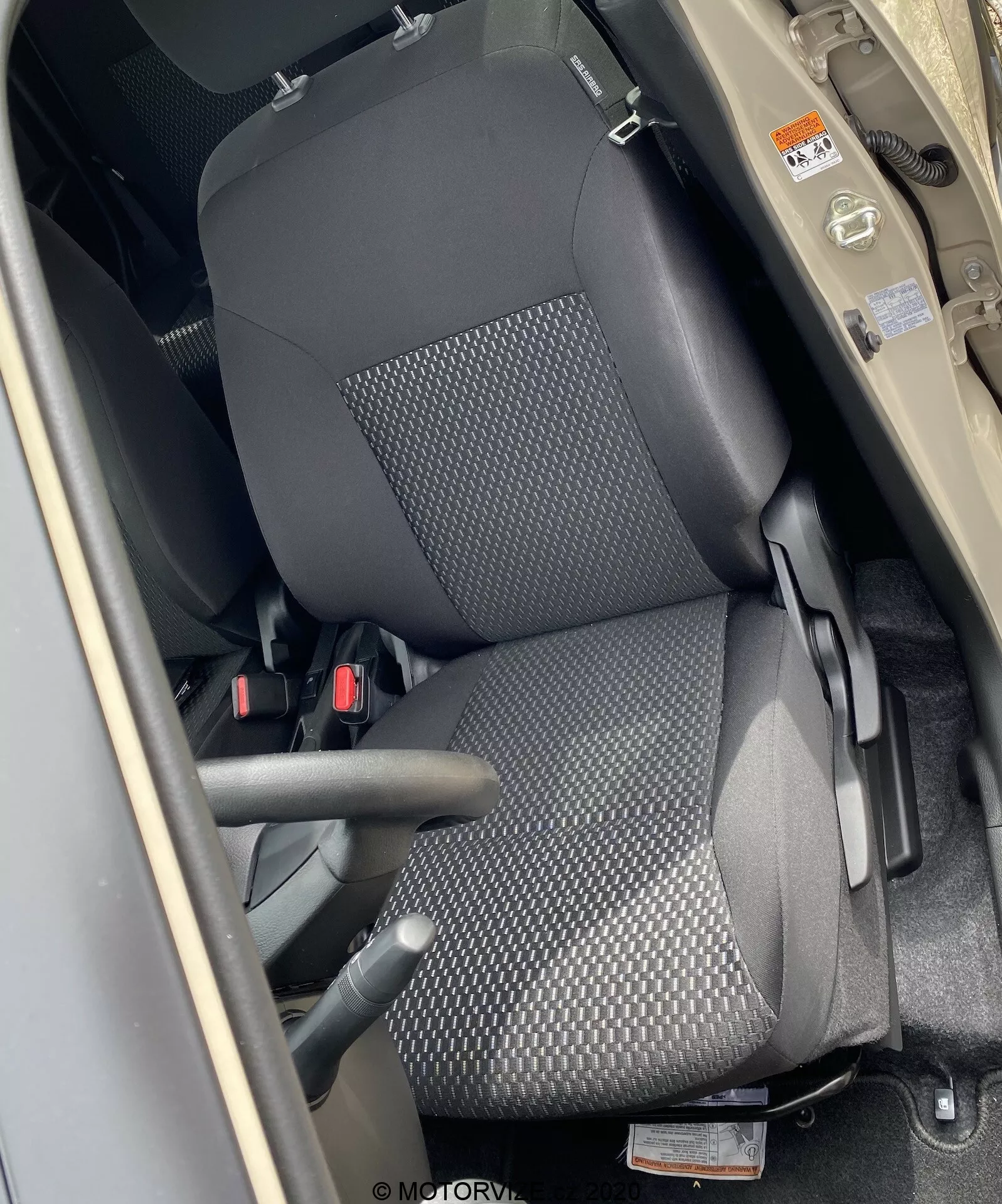 Pogled od zgoraj na sprednji sedež sovoznika v Suzuki Ignis 2020, ki prikazuje vzglavnik, naslonjalo in sedežno površino. Posnetek je narejen iz perspektive osebe, ki stoji poleg odprtih vrat, ki se nahajajo okoli stebrička B. Sedež je oblazinjen v črno tkanino z vzorcem pikčaste teksture in ima ročico za ročno nastavitev. Vidni je tritočkovni varnostni pas, notranjost v bež ali svetlo rjavi barvi pa je v kontrastu s črnim sedežem.