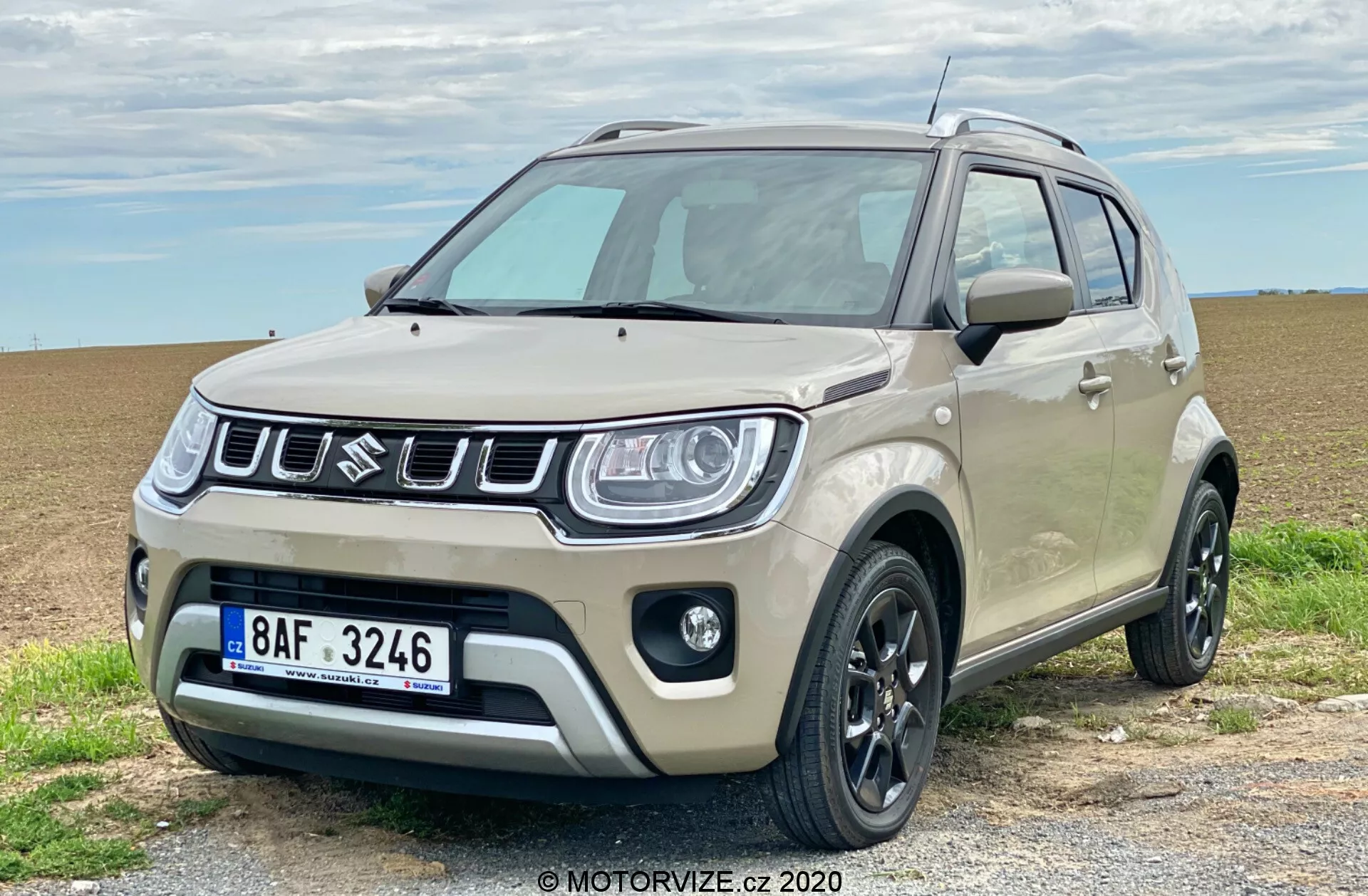 Kompaktni SUV Suzuki Ignis 1.2 Dualjet Hybrid Club 2020 je parkiran na gramozni poti z odprtim poljem v ozadju, prikazuje se v treh četrtinah sprednjega pogleda. Ima dvobarvno karoserijo, kjer je glavna barva svetlo bež, črni dodatki pa so na stranskih ogledalih, strešnih letvah in odbijačih. Značilna prednja rešetka ima navpične letvice z osrednjim simbolom znamke. Moderna žarometa z LED dnevnimi lučmi, črna lita platišča, plastična oblazinjenja, meglenke in visoka oddaljenost od tal so izrazite lastnosti. Vozilo oddaja tako urbano privlačnost kot tudi lahke terenske zmogljivosti.