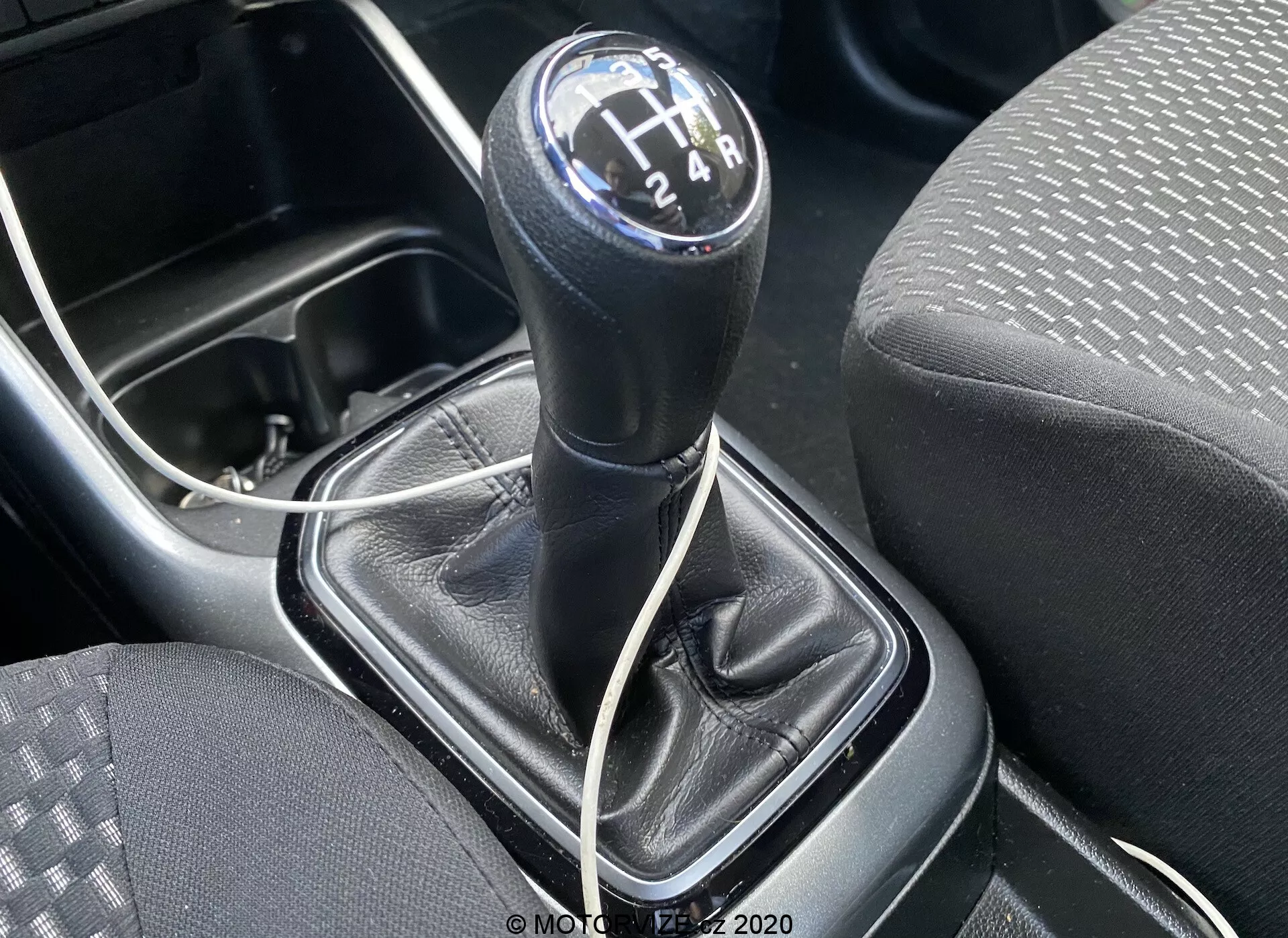 Bližnji pogled na ročico ročnega menjalnika Suzuki Ignis 1.2 Dualjet Hybrid Club 2020 iz zgornjega kota, s fokusom na okrogli črni gumb ročice z belimi številkami v 6-stopenjski H-postavitvi in vzvratno prestavo. Ročica menjalnika je v nevtralnem položaju. Okoli ročice je črna usnjena prevleka z vidnimi šivi in srebrno ali sivo plastično konzolo. Na levi strani je vidni del sedeža z blago s pikicami in črtami, in pod ročico menjalnika, delno vidni beli kabel, ki kaže na sodobne funkcije povezljivosti, kot je polnilna vtičnica. V spodnjem levem kotu je rob držala za pijačo ali majhnega shranjevalnega predala, kar poudarja praktični dizajn sredinske konzole vozila.