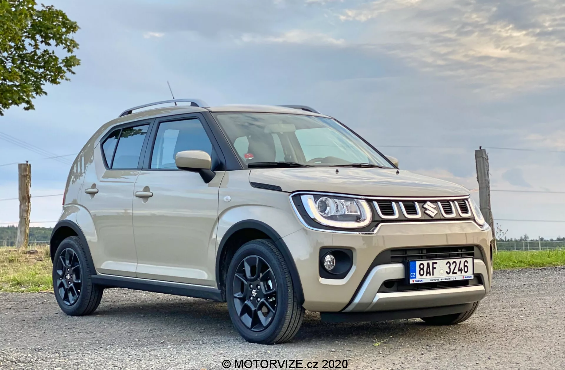 Slika prikazuje kompaktni SUV Suzuki Ignis 2020 1.2 Dualjet Hybrid Club, parkiran na ravni, verjetno asfaltirani površini, z delno oblačnim nebom v ozadju. Posneta iz nizkega kota, je sprednja stran vozila jasno vidna, kar poudarja njegovo višino in odmik od tal. Avto ima sodobno obliko s karoserijo svetle kremne ali bež barve in kontrastno črno streho. Ima črna, petkraka aluminijasta platišča z robustnimi pnevmatikami, ozke žaromete, ki segajo v blatnike, in meglenke na spodnjem odbijaču. Sprednja maska ​​ima značilno obliko z logotipom Suzuki. Stranska ogledala v dveh tonih se ujemajo s karoserijo in streho, na vrhu pa so nameščeni strešni nosilci. Tehnološke funkcije lahko vključujejo LED ali HID žaromete in dnevne luči. Spodnji del sprednjega odbijača ima robustno zasnovo s simulirano zaščitno ploščo. Na sprednji strani je vidna tudi registrska tablica. Avto zavzema velik del okvirja, kar poudarja njegov dizajn in značilnosti pri naravni dnevni svetlobi.
