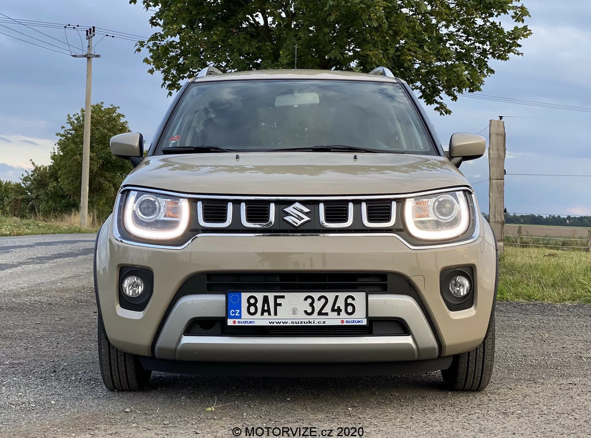 Sprednji pogled na Suzuki Ignis 2020 (Suzuki Ignis 1.2 Dualjet Hybrid Club) posnet z rahlo nizkega kota, prikazuje prednje značilnosti, vključno z rešetko z emblemom, pravokotnimi LED žarometi, meglenkami in osrednjim dovodom zraka na asfaltirani cesti z naravno krajino in oblačnim nebom v ozadju.