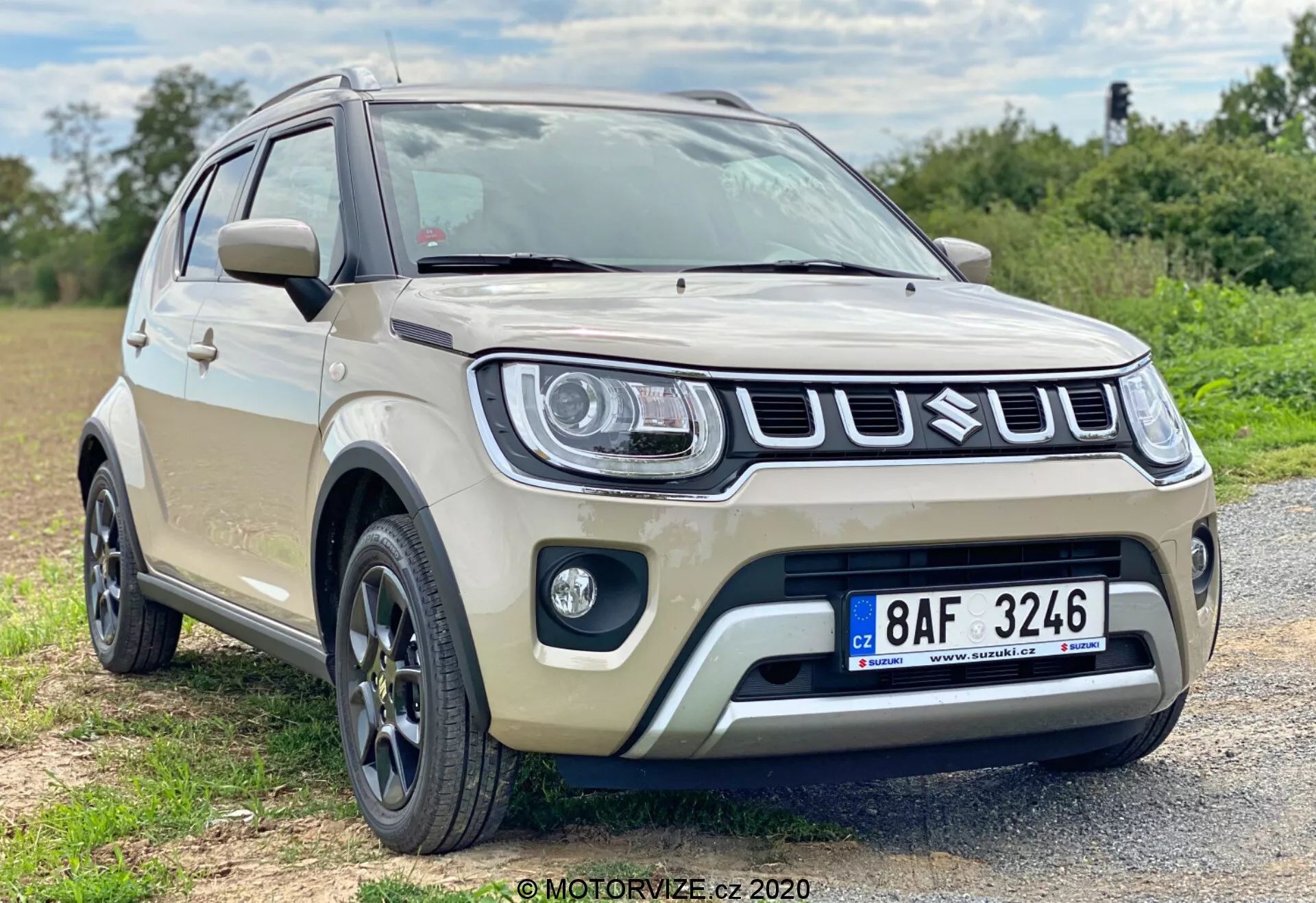 Sodobni subkompaktni crossover SUV bež barve, verjetno model Suzuki Ignis 2020, parkiran na gramozni površini na podeželskem območju. Vozilo, posneto pod nizkim kotom iz frontalnega trikotnega pogleda, prikazuje svoj značilen dizajn sprednje maske vključno z izrazito rešetko, sodobnimi žarometi in meglenkami. Platišča iz lahke kovine z večkrakim dizajnom, višja oddaljenost od tal, zaščitne obloge in bočna ogledala z integriranimi smerniki poudarjajo njegove sodobne in robustne značilnosti. Plitva globinska ostrina z naravno osvetlitvijo se osredotoča na vozilo.