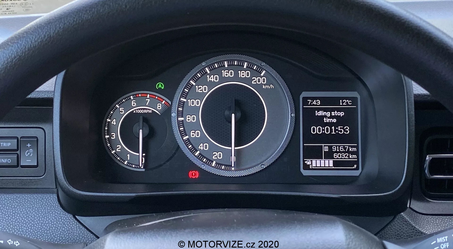 Posnetek notranjosti Suzuki Ignis 2020 osredotočen na instrumentno ploščo za volanskim obročem. Vidni sta zgornji del volanskega obroča in krmilni ročici za smerne utripalke in brisalce. Instrumentna plošča združuje analogne in digitalne prikazovalnike: na levi strani je analogni merilnik vrtljajev z rdečim poljem pri 6500 RPM, na desni strani pa analogni merilnik hitrosti do 220 km/h. Centralni digitalni prikazovalnik prikazuje zunanjo temperaturo (12°C), čas (7:43), funkcijo 'Idling stop', časovnik (00:01:53) in prevožene kilometre (91672 km). Moderen, uporabniku prijazen dizajn značilen za tržišča z metričnimi merskimi enotami.