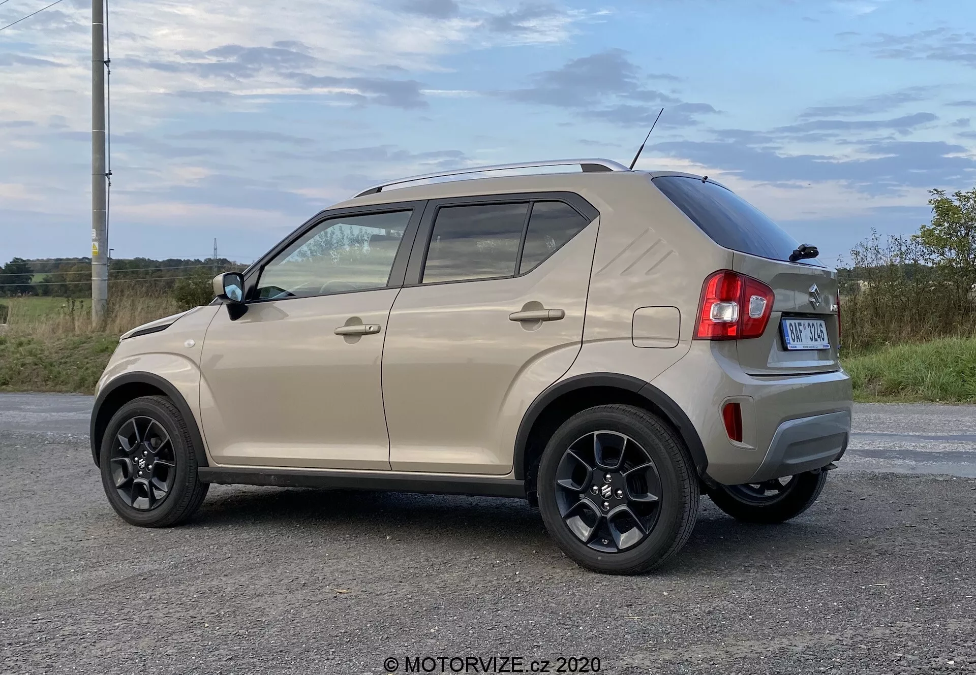 Kompaktni SUV Suzuki Ignis 1.2 DualJet Hybrid Club 2020 v zadnjem kotu s treh četrtin, s hrbtnim pokrovom z zadnjim brisalcem, aerodinamično strešno linijo, visoko nameščeno zavorno lučjo in anteno morskega psa. Stranski prikaz kaže črna večkraka platišča, barvne ročaje vrat z vgrajenimi smerniki, plastične zaščite na spodnjem delu vrat in stranskih pragov ter črni okvir okoli oken. Vozilo je parkirano v naravnem podeželskem okolju na asfaltni površini podnevi.