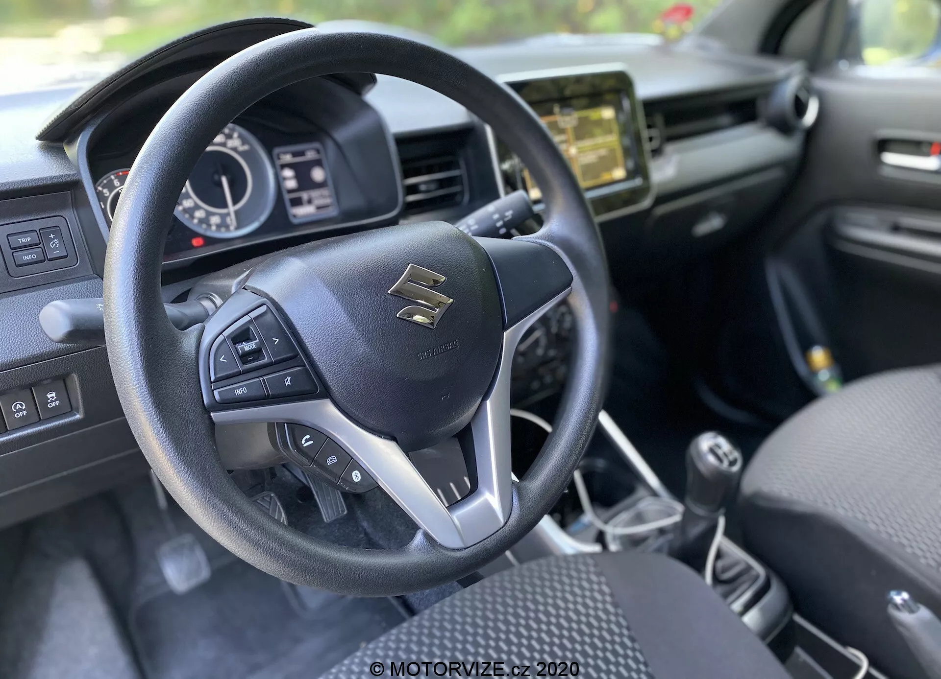 Pogled na notranjost iz voznikove perspektive v Suzuki Ignis 2020 (1.2 Dualjet Hybrid Club) s poudarkom na sodobnem trikrakam volanu z večfunkcijskimi gumbi. Suzuki emblem je na sredini, obdano s srebrnimi poudarki in kakovostnim šivanjem. Za volanom je deloma viden instrumentni zaslon z digitalnim prikazom in analognimi kazalci, ki prikazujejo hitrost, obrate in stanje vozila. Sredinska konzola ima informacijsko-razvedrilni sistem z zaslonom, ki verjetno ponuja avdio, navigacijo in nastavitve vozila. Sredinska konzola ima ročico ročnega menjalnika, kar nakazuje na ročni menjalnik. Notranja oprema je pretežno črna s teksturnimi elementi, sedeži z vzorčnim dizajnom, kar nakazuje na estetiko in udobje.