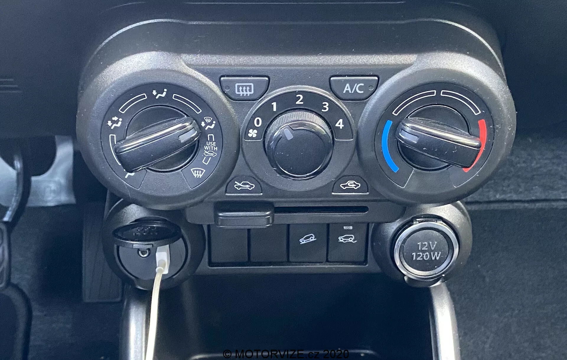 Od blizu centeralne konzole Suzuki Ignis 2020, osredotočen na enoto za nadzor klime s tremi glavnimi gumbi za temperaturo, hitrost ventilatorja in smer pretoka zraka. Gumb za temperaturo ima modre in rdeče indikatorje za hladnejše in toplejše nastavitve. Nad osrednjim gumbom za hitrost ventilatorja je gumb 'A/C', gumb za recirkulacijo zraka v kabini in gumbi za prednje odroševanje. Pod kontrolami je vtičnica 12V 120W, kar poudarja praktičen in uporabniku prijazen dizajn vozila z ročnimi stikali.