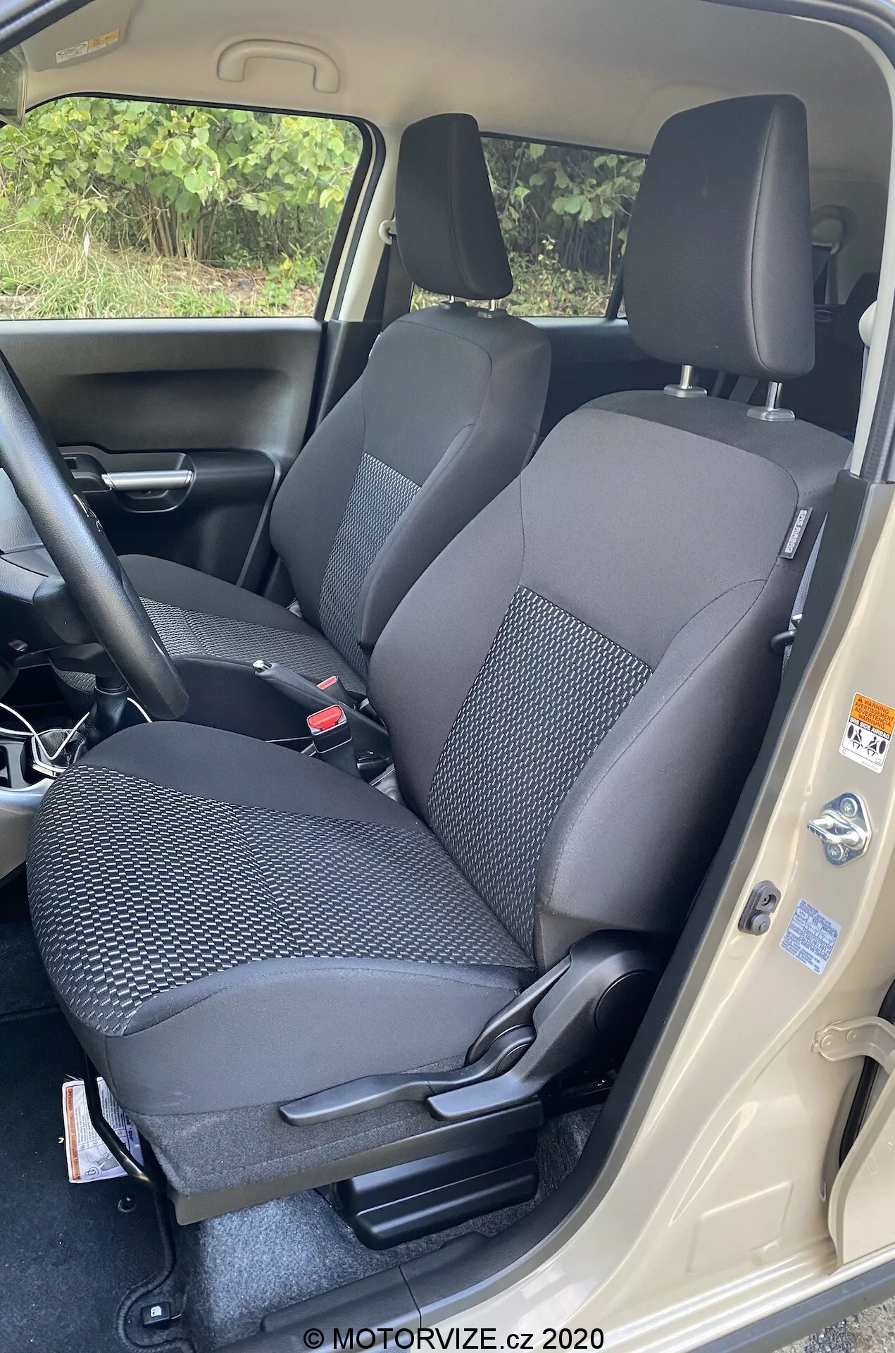 Profesionalna fotografija notranjosti Suzuki Ignis 2020 (Suzuki Ignis 1.2 Dualjet Hybrid Club), osredotočena na območje sedeža sprednjega potnika. Posneta z visoke perspektive, kamera zajame jasen pogled z odprtimi vrati avtomobila. Dvobarvna tkaninska oblazinjenje sedeža ima vzorčast dizajn za trajnost. Nastavitve sedeža so ročne, z naslonom za glavo, varnostnim pasom in vidnimi ročicami za prilagoditev. Vidna je tudi del sredinske konzole in notranja plošča vrat z oznakami za regulacijo. Temno preprogo in svetel prag vrat poudarjata kontrast dizajna. Slika, pripisana MOTORVIZE.cz 2020.
