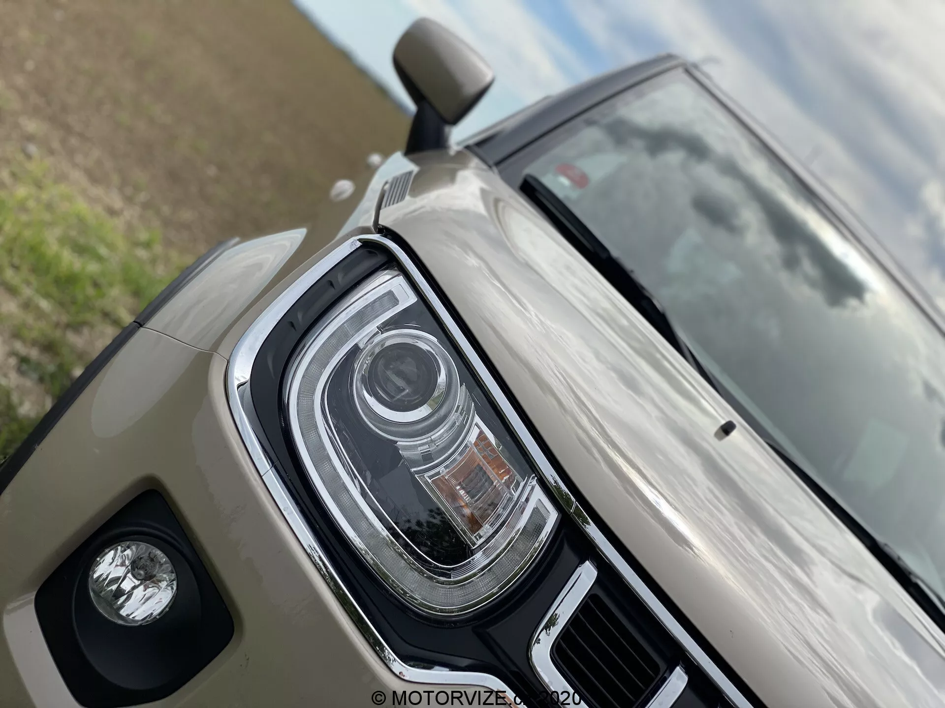 Podroben pogled Suzuki Ignis 2020, poudarjena leva sprednja kvadranta. Značilnosti LED žarometov z več oddelki, verjetno prilagodljivi žarometi ali samodejni dolgi žarometi. Prikazuje svetlo bež/svetlo sivo sijočo površino in ohišje meglenke. Delni pogled na stransko ogledalo namiguje na napredno tehnologijo, kot so smerniki na stranskih ogledalih ali nadzor mrtvih kotov.