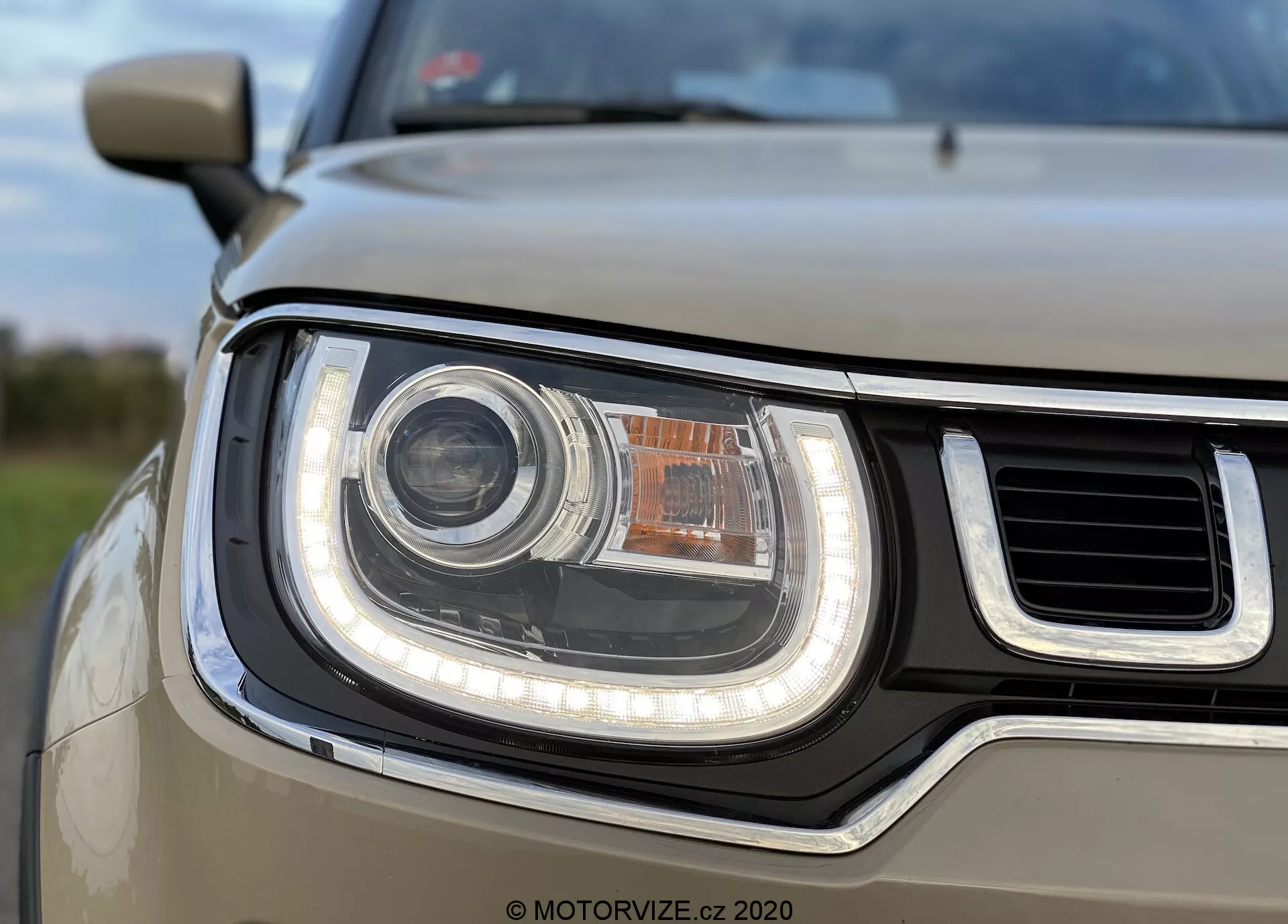 Povečan pogled na sklop žarometov na sprednji levi strani Suzuki Ignis 2020, ki prikazuje verjetno LED ali HID krožni glavni žaromet, obdan z LED dnevno lučjo (DRL) v obliki cevi. Žaromet ima zgoraj manjšo, morda oranžno smerno ali parkirno svetilko. Delno vidna mreža ima vodoravne palice, zasnova žarometa pa se z ukrivljenimi in ravnimi robovi daje moderen in robusten videz. Globinska ostrina je osredotočena na žaromet, medtem ko so ozadje in drugi deli avtomobila nekoliko zamegljeni.