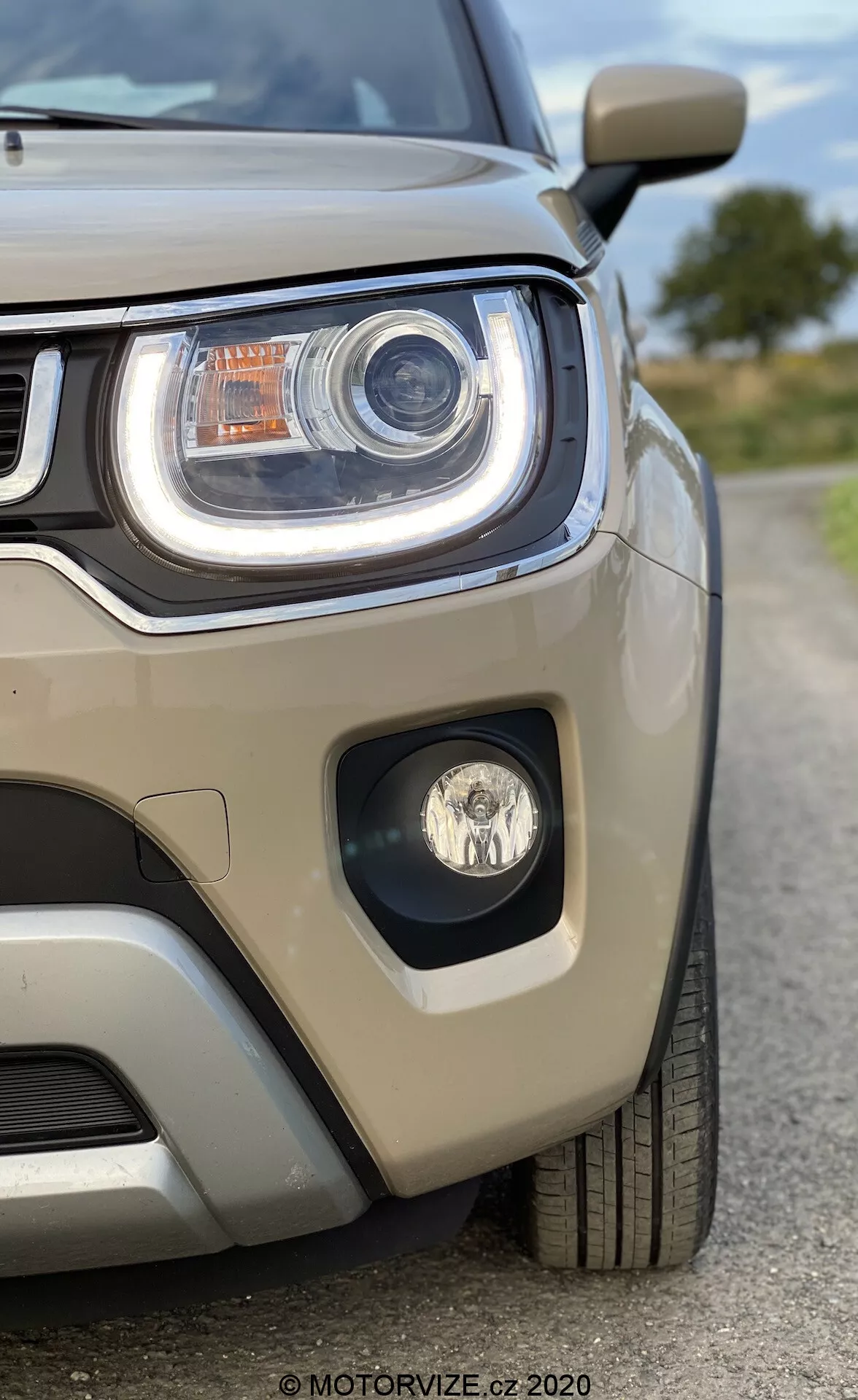 S pogledom s sprednje leve strani Suzuki Ignis 2020 s poudarkom na sklopu žarometa in delu rešetke. Slika, posneta iz rahlo nizkega kota, poudarja agresivne linije žarometa. Odlikuje ga izrazita LED dnevna luč, projektorska tehnologija, dodatni svetlobni elementi in meglenka v ločenem okviru. Svetlo bež kovinska barva odseva svetlobo, vidno parkirno tipalo na odbijaču pa poudarja parkirno asistenco. Avto deluje zelo čisto in dobro vzdrževano, kar pritegne pozornost na njegov sodobni dizajn.