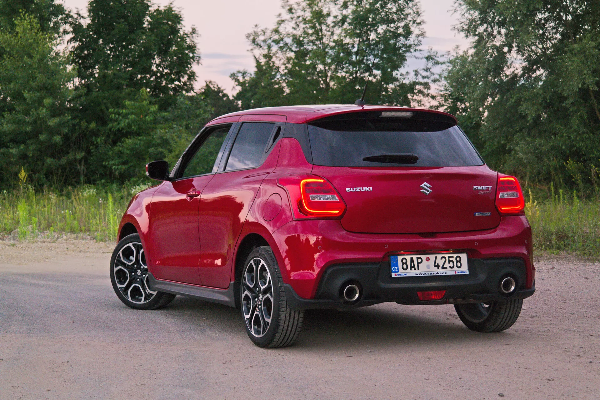 Slika rdečega Suzuki Swift 2017 Swift-Sport v pogledu tri četrt od zadaj, ki prikazuje sodobne oblikovne lastnosti, kot so LED zadnje luči, integriran spojler, antena v obliki plavuti morskega psa, večkrake aluminijaste platišča, kljuke vrat in stranska ogledala v barvi karoserije ter oznaka hibridne tehnologije. Naravno ozadje dodaja občutek miru in dinamike.
