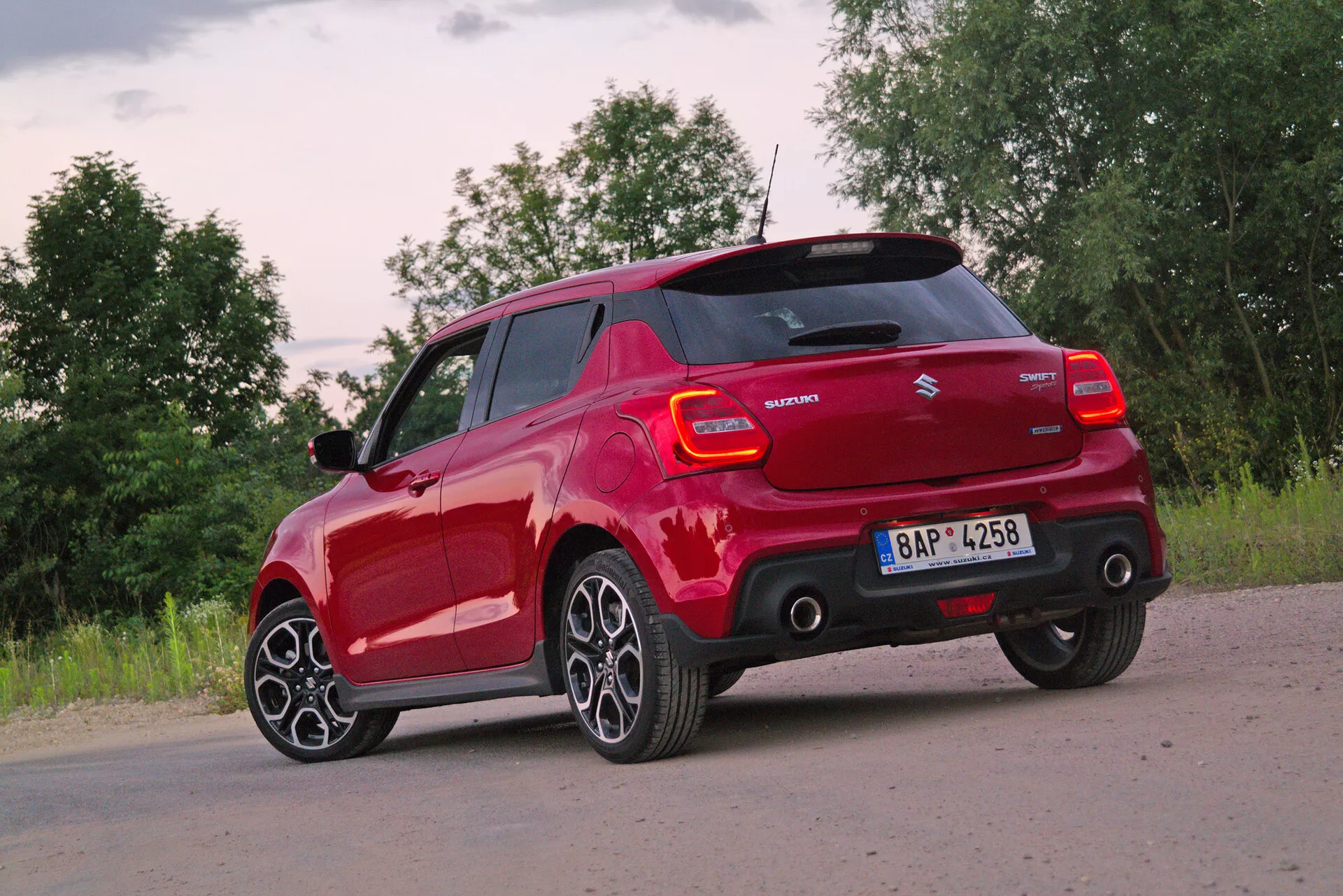Rdeči Suzuki Swift Sport 2017 hatchback zajet z zadnjega trikotnega kota, poudarjajoč zadnji in bočni profil na voznikovi strani. Fotografija, posneta iz nekoliko znižane perspektive, poudarja moderne večkrake aluminijaste platišča in pnevmatike z nizkim profilom. Osvetlitev ob zori ali mraku z delno oblačnim vremenskim stanjem omogoča mehko naravno svetlobo. Vidne funkcije vključujejo sodoben dizajn zadnjih luči z LED elementi, značko proizvajalca, ime modela, stranska ogledala v barvi karoserije, morda s smerokazi, zadnji odbijač z integriranimi parkirnimi senzorji in strešno linijo, ki nakazuje na spojler za aerodinamično učinkovitost.