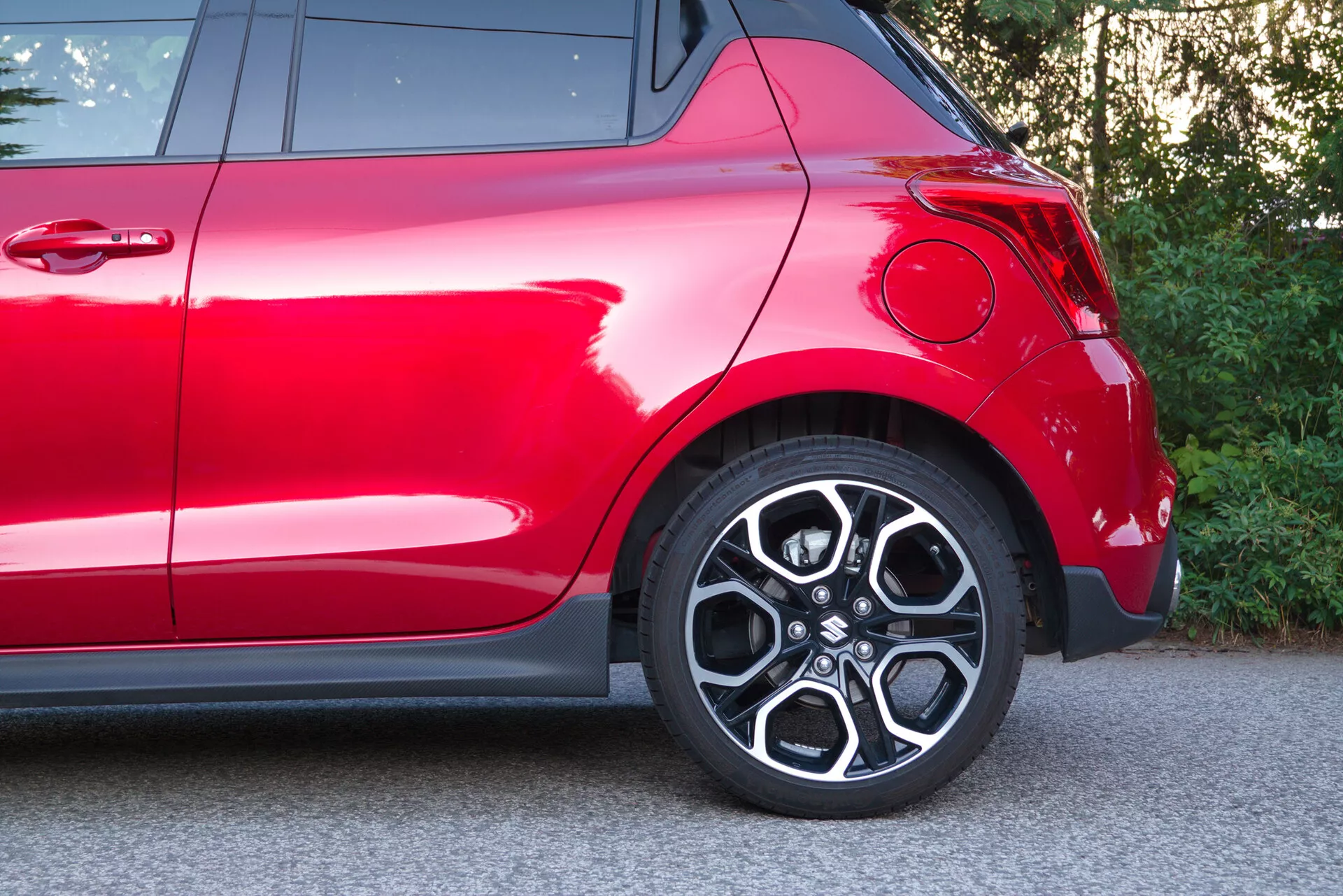 Delni bočni pogled na rdeči Suzuki Swift 2017 (Swift Sport), osredotočen na zadnji del, vključno z zadnjim kolesom in območjem blatnika. Bližnji posnetek zajema elegantno lito platišče z večkrakim dizajnom, pnevmatiko z nizkim profilom, moderen ukrivljen zadnji blatnik, globoko sijočo rdečo barvo, del zadnje luči in kromirane okrasne elemente, ki poudarjajo njegov športni dizajn in vrhunsko zunanje zaključke.
