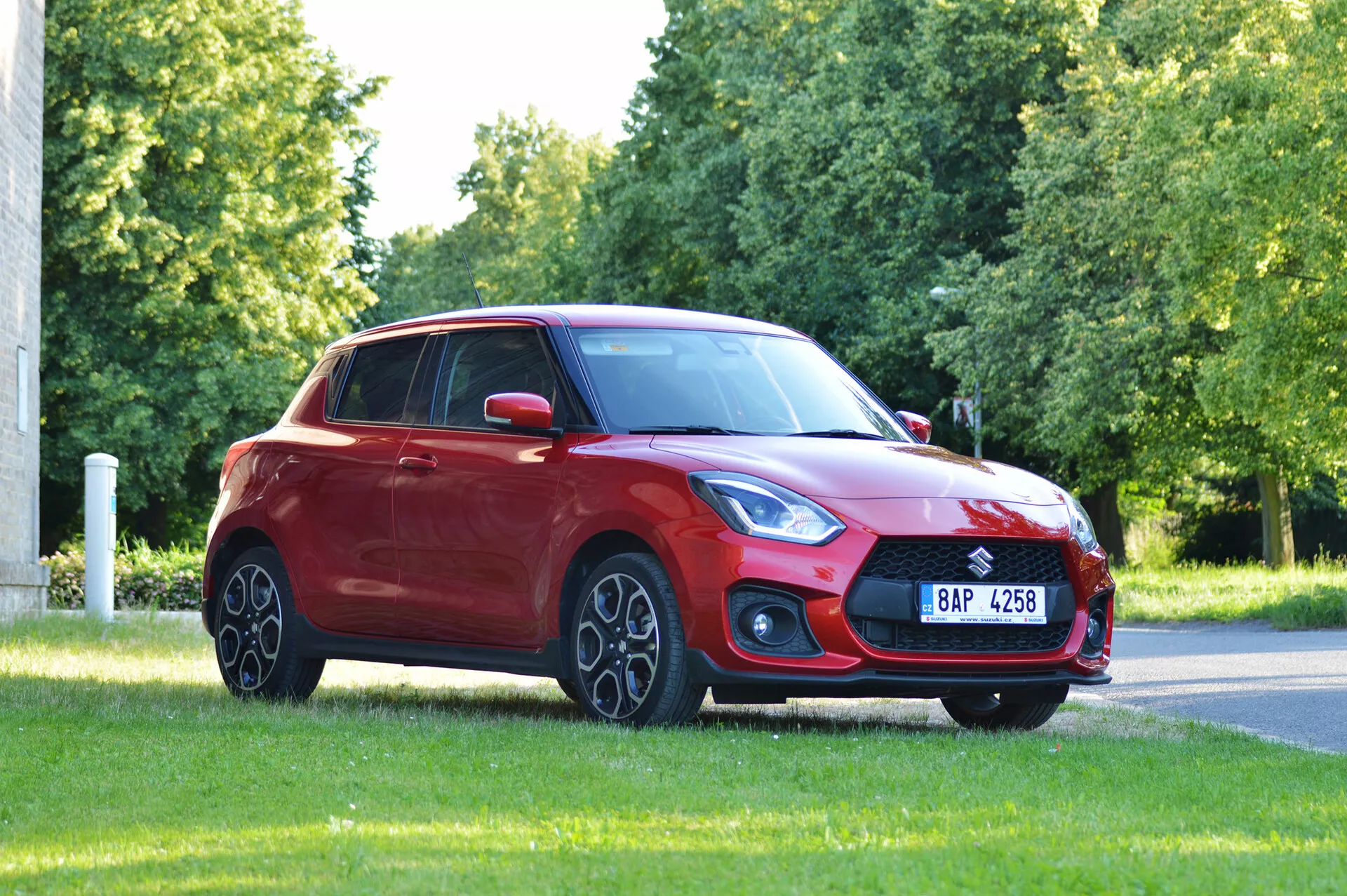 Slika prikazuje rdeč hatchback Suzuki Swift Sport iz leta 2017 na travnati površini, verjetno v parku ali ob cesti, posnet iz sprednjega tripetinskega kota. Izstopajo izrazita sprednja rešetka, elegantne in kotne žaromete, sestav meglenk in nagnjena pokrov motorja v aerodinamično vetrobransko steklo. Lita platišča, stranska ogledala v barvi karoserije, nežno padajoča linija strehe proti zadnjemu oknu in zatemnjena okna prispevajo k dizajnu vozila. Naravna osvetlitev in neoviran pogled poudarjata celotno držo in oblikovanje avtomobila.