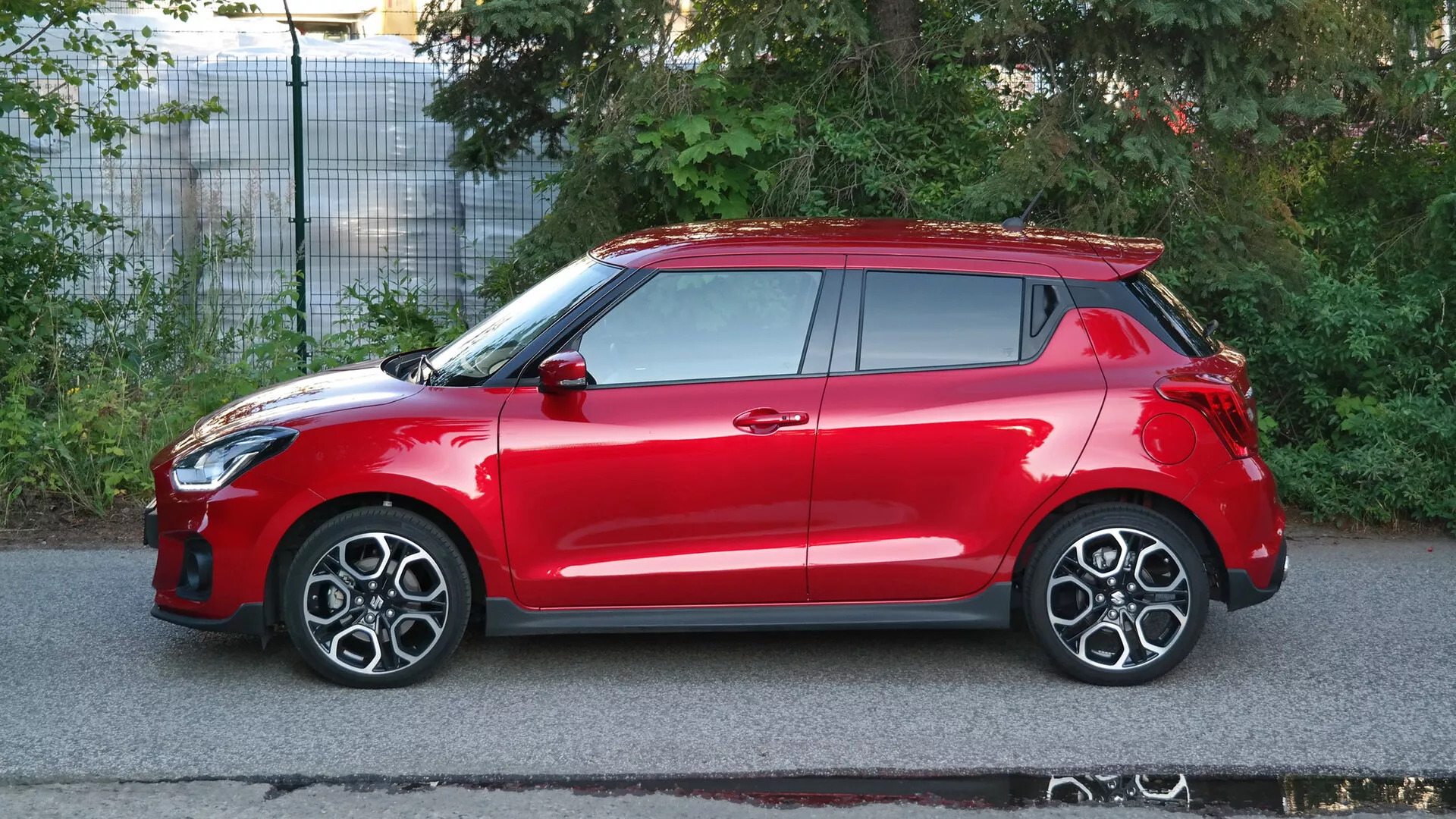 Stranski profil rdečega hatchback avtomobila Suzuki Swift Sport 2017. Vozilo je posneto v naravni dnevni svetlobi, kar poudarja elegantne oblikovne značilnosti, vključno z integracijo žarometov v sprednji obroč kolesa, večkrakimi litimi platišči, stranskimi ogledali in kljukami v barvi karoserije, sistemom za brezključni vstop in vidno strešno okno. Avto ima tudi strešni spojler in visoko nameščene zadnje luči. Posnetek, narejen v predmestnem okolju, poudarja aerodinamičnost avta, njegove športne značilnosti in verjetno napredne varnostne tehnologije.