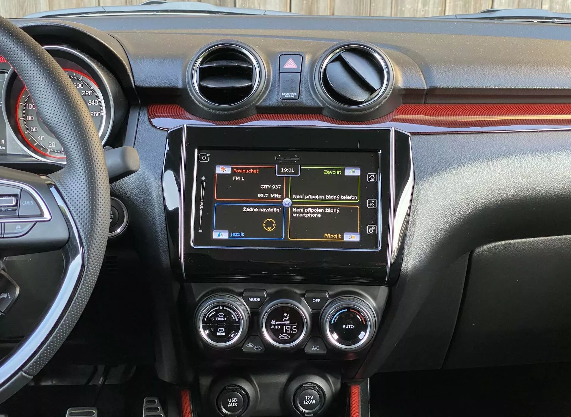 Podroben pogled na notranjost Suzukija Swift Sport 2017, osredotočen na osrednjo nadzorno ploščo iz perspektive zadnjega sedeža, prikazuje modernega infotainment sistema, okrogle zračnike klimatske naprave, HVAC kontrole, različne funkcijske gumbe, dvobarvno oblikovanje nadzorne plošče ter del volana z nameščenimi kontrolami.