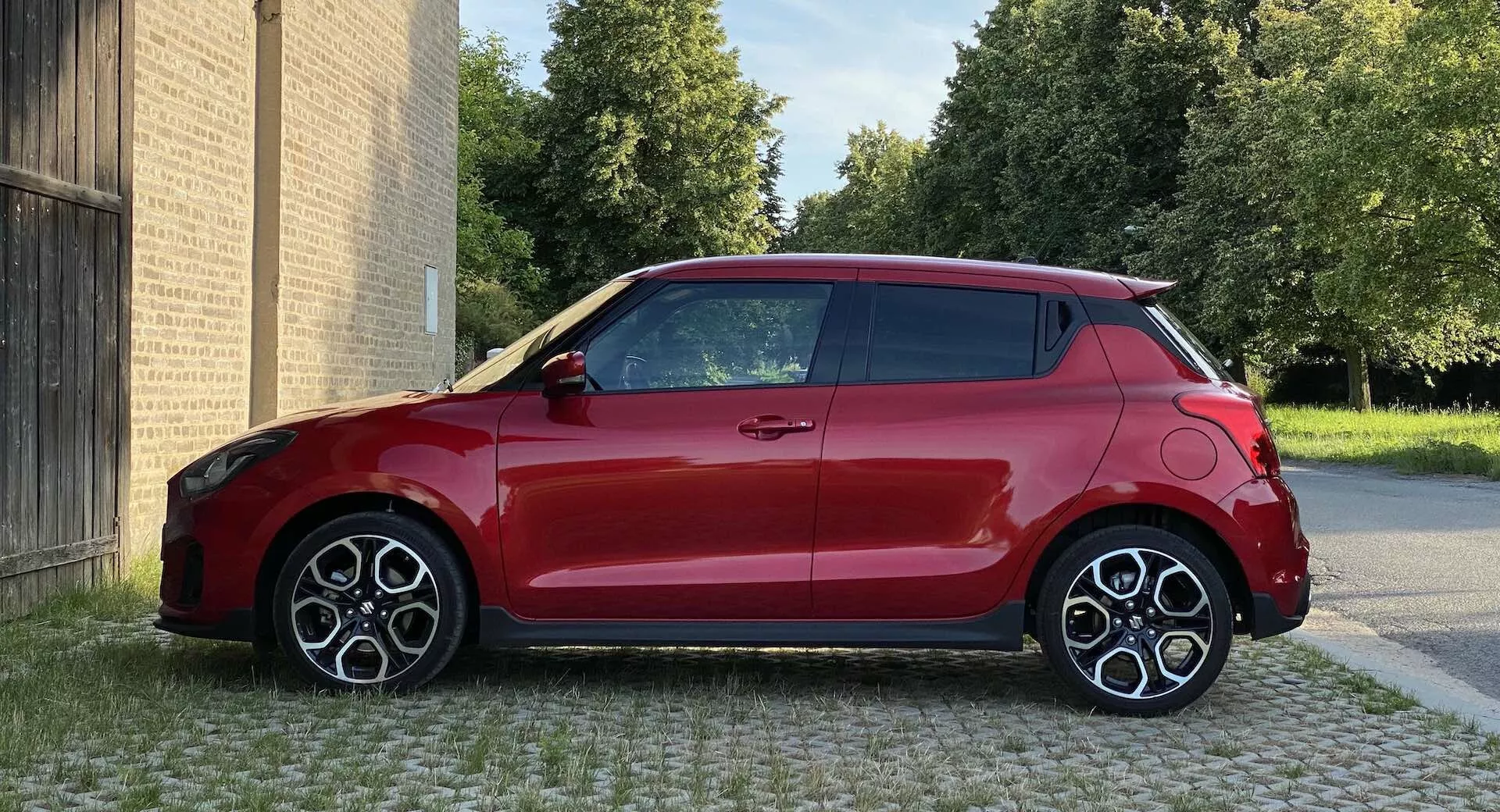 Slika rdečega Suzuki Swift Sport hatchbacka iz leta 2017, posneta z bočnega profila na višini oči, prikazuje desno stran z dvobarvno zasnovo rdeče karoserije in črne strehe. Avto je parkiran na tlakovanem območju, v ozadju so zgradbe, zadaj pa so vidna drevesa. Kompaktna, moderna zasnova ima velika športna večkraka platišča, vidne aerodinamične linije in anteno v obliki plavuti morskega psa. Osvetlitev je dnevna, kakovost slike je jasna in vidni so površinski odsevi.