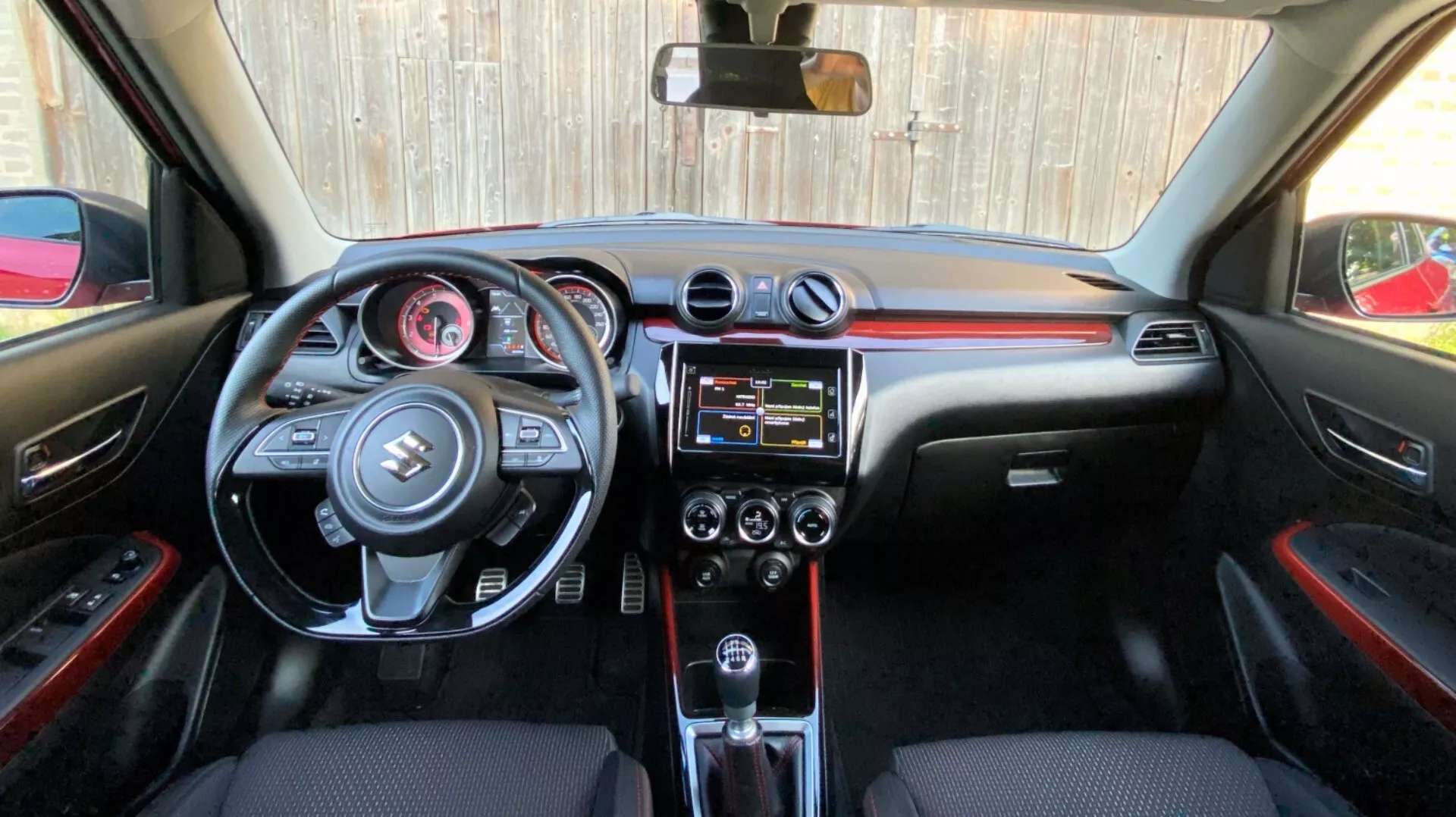 Notranji pogled Suzuki Swift 2017 (Swift Sport) s perspektive zadnjega sedeža, usmerjen proti sprednji kabini, prikazuje podroben pogled na armaturno ploščo, volan z vgrajenimi kontrolami, osrednjo konzolo s sistemom za informiranje in zabavo ter nadzorom klimatske naprave, prestavno ročico avtomatskega menjalnika in črne notranje materiale z rdečimi poudarki.