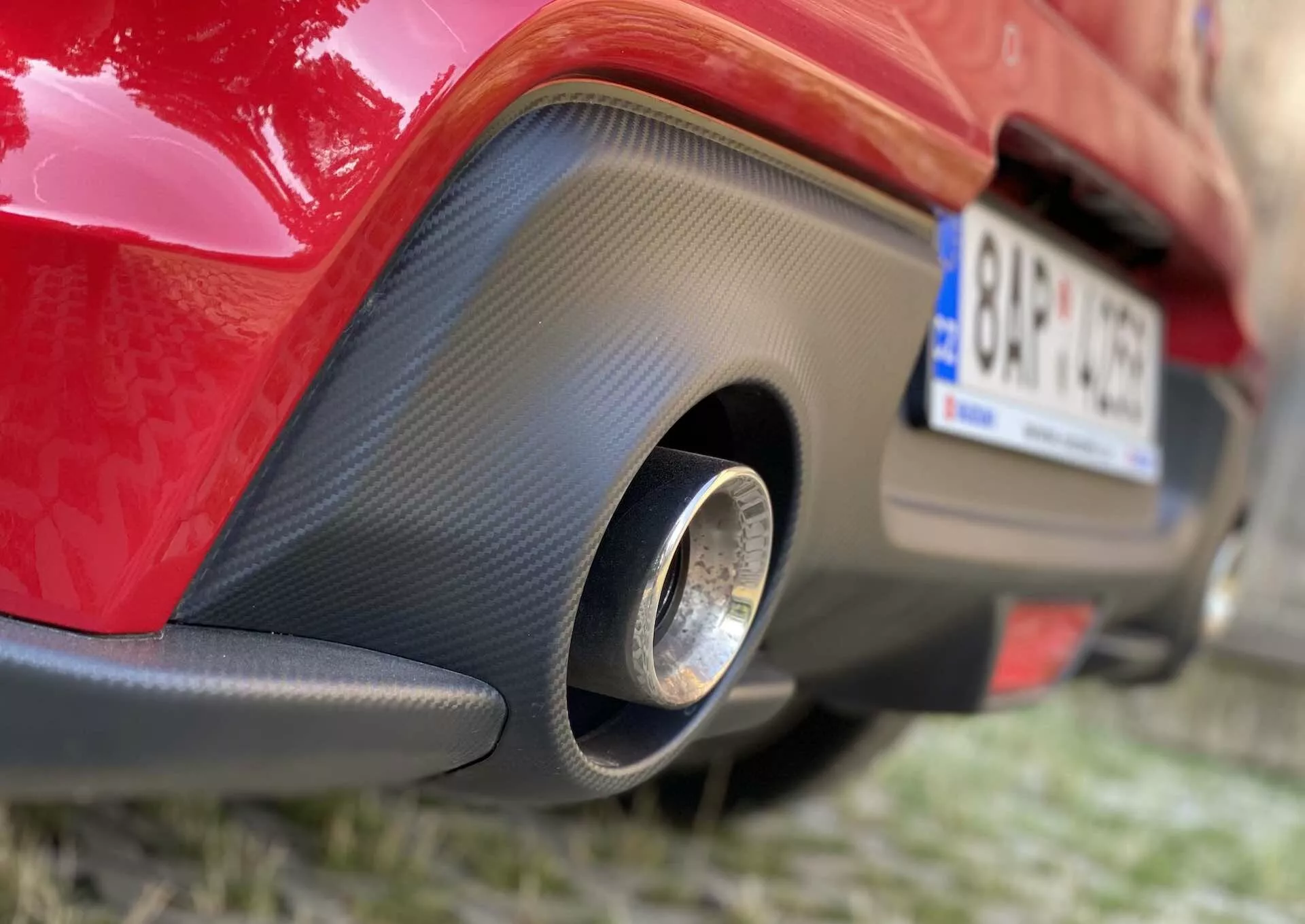 Posnetek od blizu zadnjega dela rdečega Suzuki Swift Sporta iz leta 2017, ki se osredotoča na spodnji del zadnjega odbijača. Fotografija zajame nižen kot z desne strani, poudarjajoč črn vložek, podoben karbonskim vlaknom, s posamezno okroglo kovinsko izpušno cevjo na sredini.