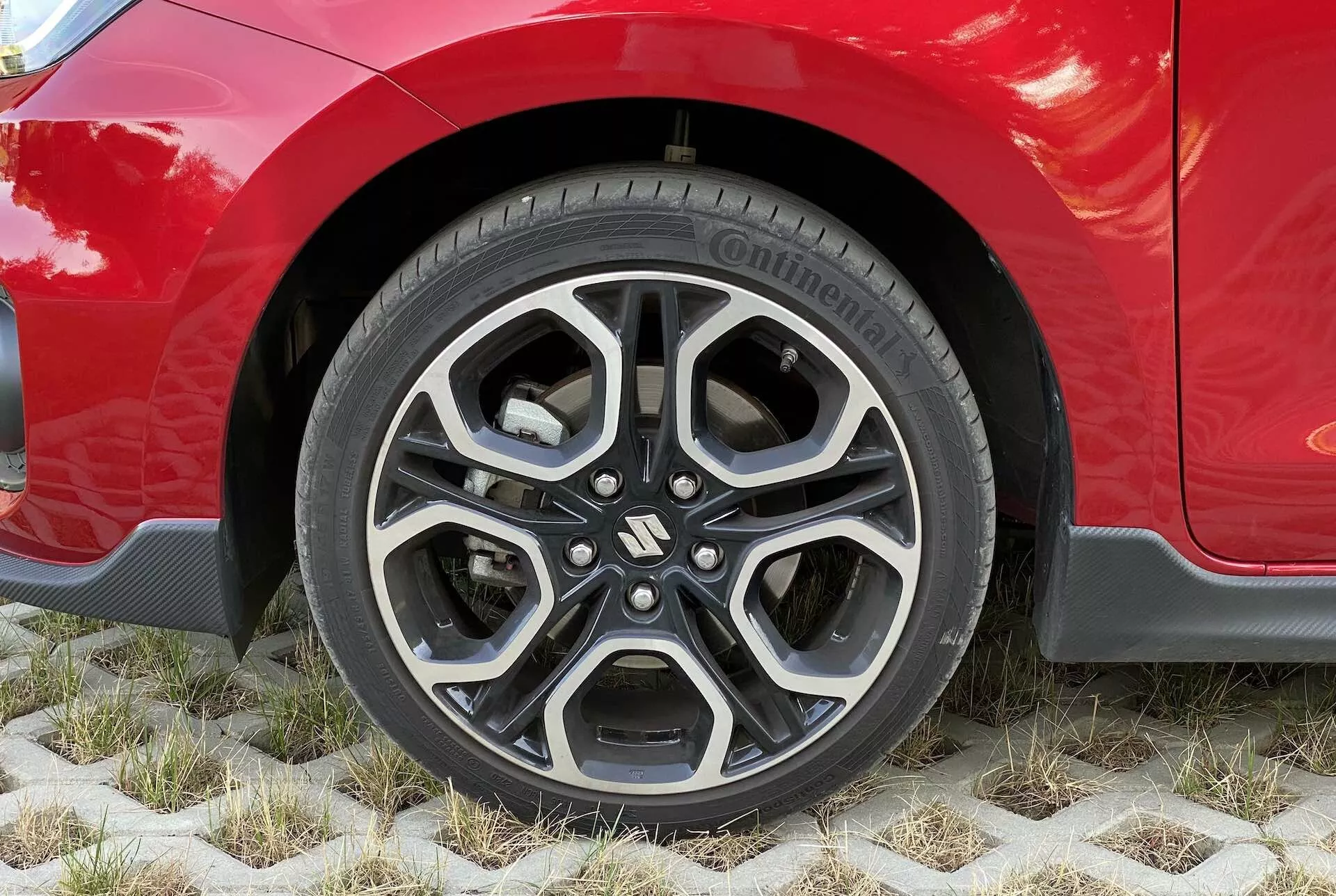 Približan posnetek kolesa rdečega Suzuki Swift 2017 Swift Sport, ki prikazuje moderno aluminijasto platišče z zapletenim, večkrakim dizajnom v črni in obdelani srebrni barvi. Vidna pnevmatika je model Continental PremiumContact, kar kaže na visoko kakovost in dobro stanje pnevmatike. Karoserija avtomobila, svetleča in dobro vzdrževana, sega nad kolo, poudarjajoč estetske linije blatnika in morda športno opremo. Vozilo je parkirano na ekoloških šesterokotnih tlakovanih površinah, ki omogočajo rast trave, kar poudarja trajnostne rešitve za parkiranje.