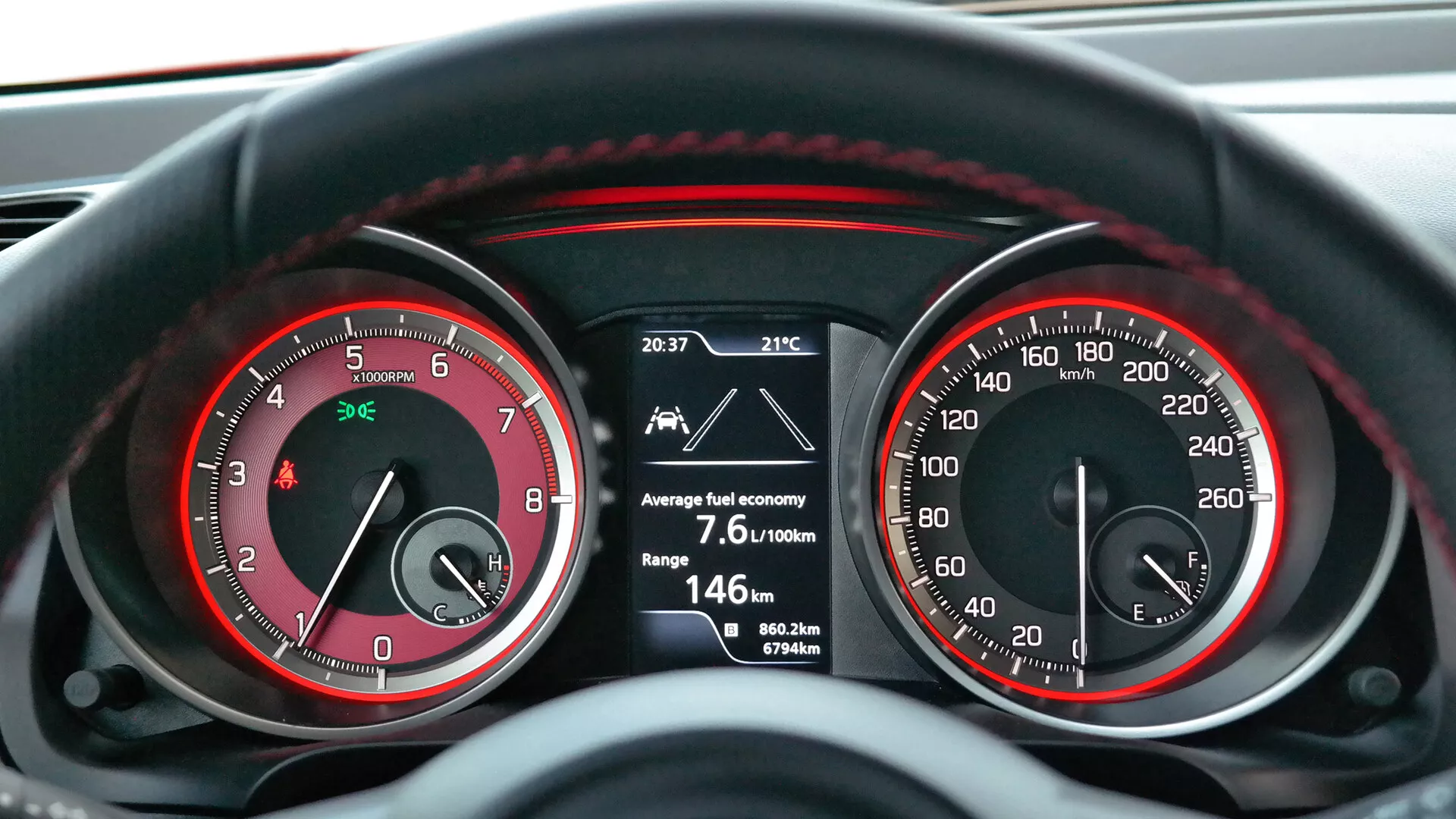 Od blizu, z vidika voznika prikazuje sliko armaturne plošče v Suzuki Swift 2017 (Sport) z analognim merilnikom vrtljajev (do 7 000 RPM) in merilnikom hitrosti (do 260 km/h) z rdečimi poudarki ter osrednji digitalni zaslon, ki prikazuje čas 20:37, zunanjo temperaturo 21°C, povprečno porabo goriva 7,6L/100km, preostali doseg 146 km, dva potovalna števca (0,0 km in 367,6 km) ter skupno število prevoženih kilometrov 80.622 km. Osvetlitev nakazuje, da je vozilo v delovanju, verjetno v slabi svetlobi, kar odraža kombinacijo analogne in digitalne armaturne tehnologije.