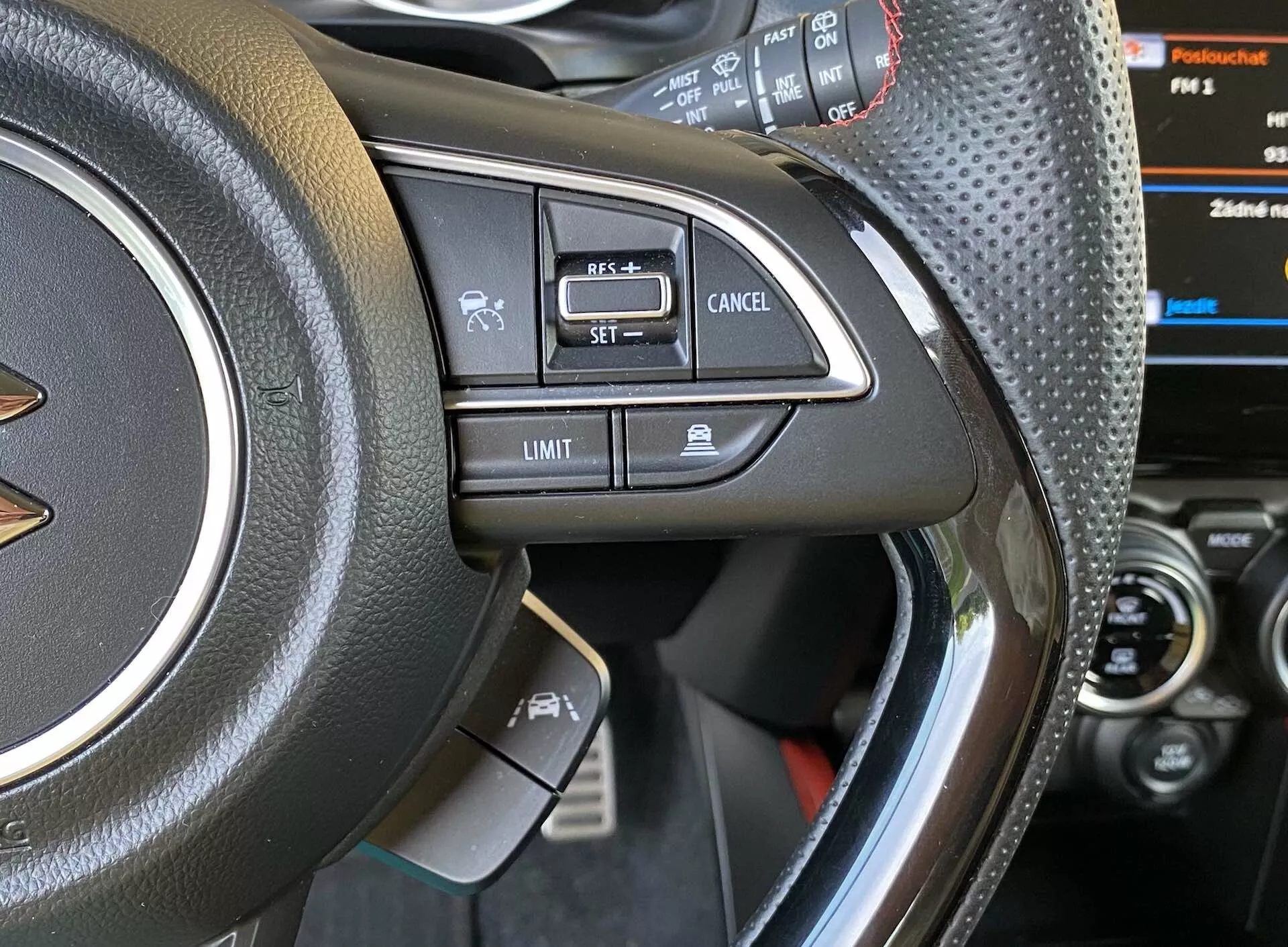 Bližnji pogled na levi del volana Suzuki Swift 2017 Swift-Sport z osredotočenjem na nameščene krmilnike. Fotografija posneta s sedeža voznika ali rahlo zunaj voznikovih vrat, osredotočena na usnjen ali usnju podoben material z vidnimi šivi, kovinski poudarek in različne gumbe, kot so glasovno upravljanje, Bluetooth prostoročna klicna funkcija, tempomat 'SET,' 'Cancel' in omejevalnik hitrosti 'Limit.' Delno viden instrumentni zaslon z digitalnim prikazom in osrednjo konzolo vozila.