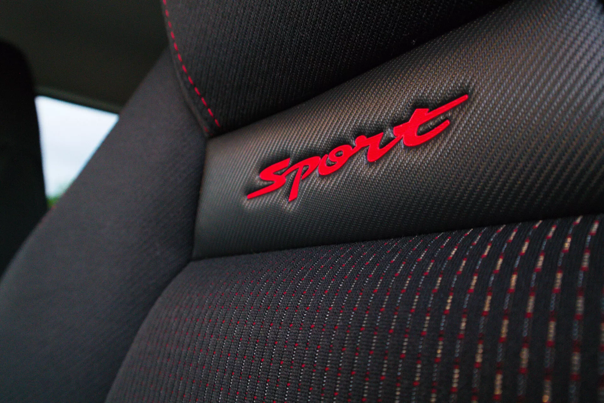 Povečava posnetek naslonjala sedeža vozila Suzuki Swift Sport 2017, ki prikazuje podrobno teksturo blaga, rdeče šive in vezenje 'Sport' na črnem blagu, kar poudarja športno opremo vozila.