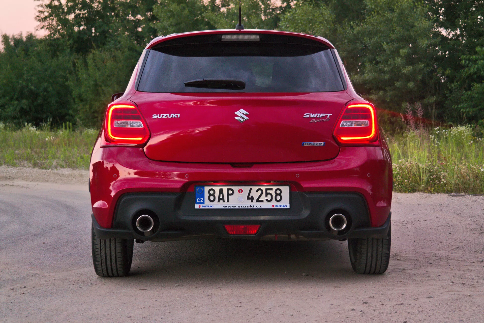 Zadnji pogled rdečega avtomobila Suzuki Swift Sport 2017, osredotočen na celotno širino in izrazit zadnji odbijač, značilne zadnje svetlobne sklope, strešni spojler, osrednji znak Suzuki, ime modela 'SWIFT', vidno izpušno cev in registrsko tablico v podeželskem okolju s zelenjem in drevesi.