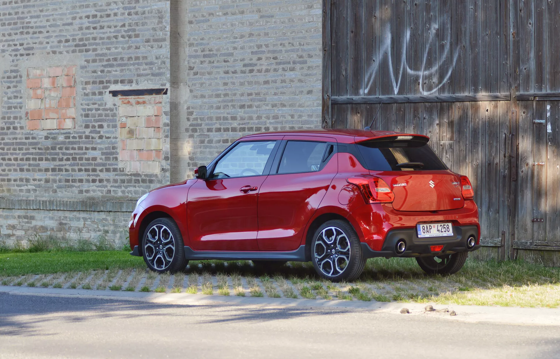 Suzuki Swift Sport hatchback 2017 v svetli rdeči barvi, posnet z zadnjega tričetrtinskega pogleda, parkiran pred staro stavbo. Slika poudarja oblikovne značilnosti, kot so zadnje luči, zadnja vrata, izrazit zadnji spojler, dvobarvni odbijač z difuzorjem, en sam vidni izpuh, večkraki lita platišča, popolnoma pobarvana karoserija, vključno s stranskimi ogledali in kljukami vrat, kar nakazuje višjo raven opreme ali luksuzne možnosti.