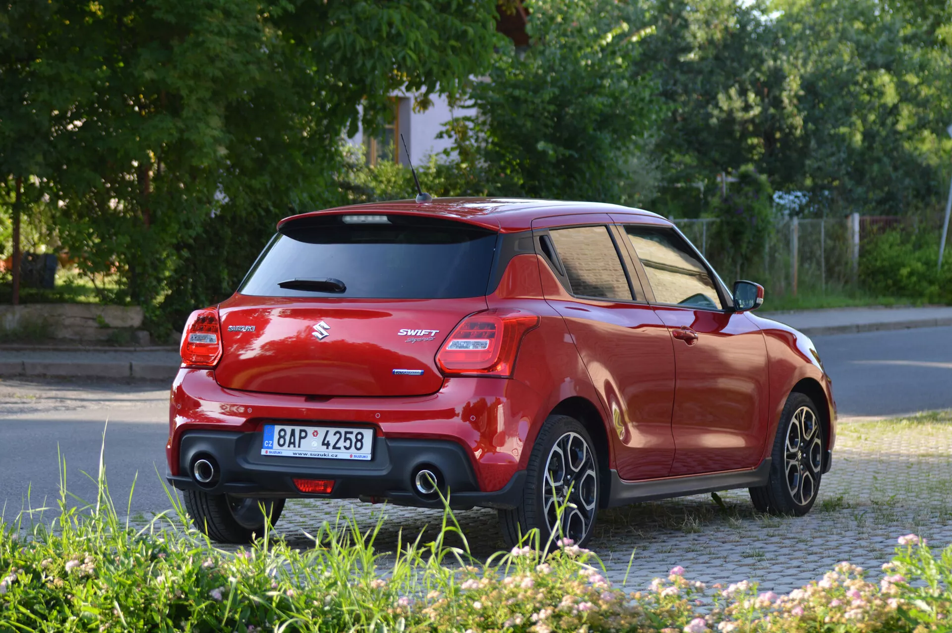 Slika prikazuje moderen hatchback Suzuki Swift Sport 2017, ujet iz zadnjega tretjinskega kota, ki prikazuje stran in zadnji del vozila. Telo izstopa s kontrastno črno streho in stebrički, dvignjenim zadnjim spojlerjem, značilnimi zadnjimi lučmi in modelno oznako 'Swift'. Fotografija je posneta na nivoju tal, prikazuje petkrake aluminijaste platišča v gibanju, kar nakazuje, da je vozilo v vožnji. Osvetlitev nakazuje, da je zgodnje jutro ali pozno popoldne, z dolgimi sencami.