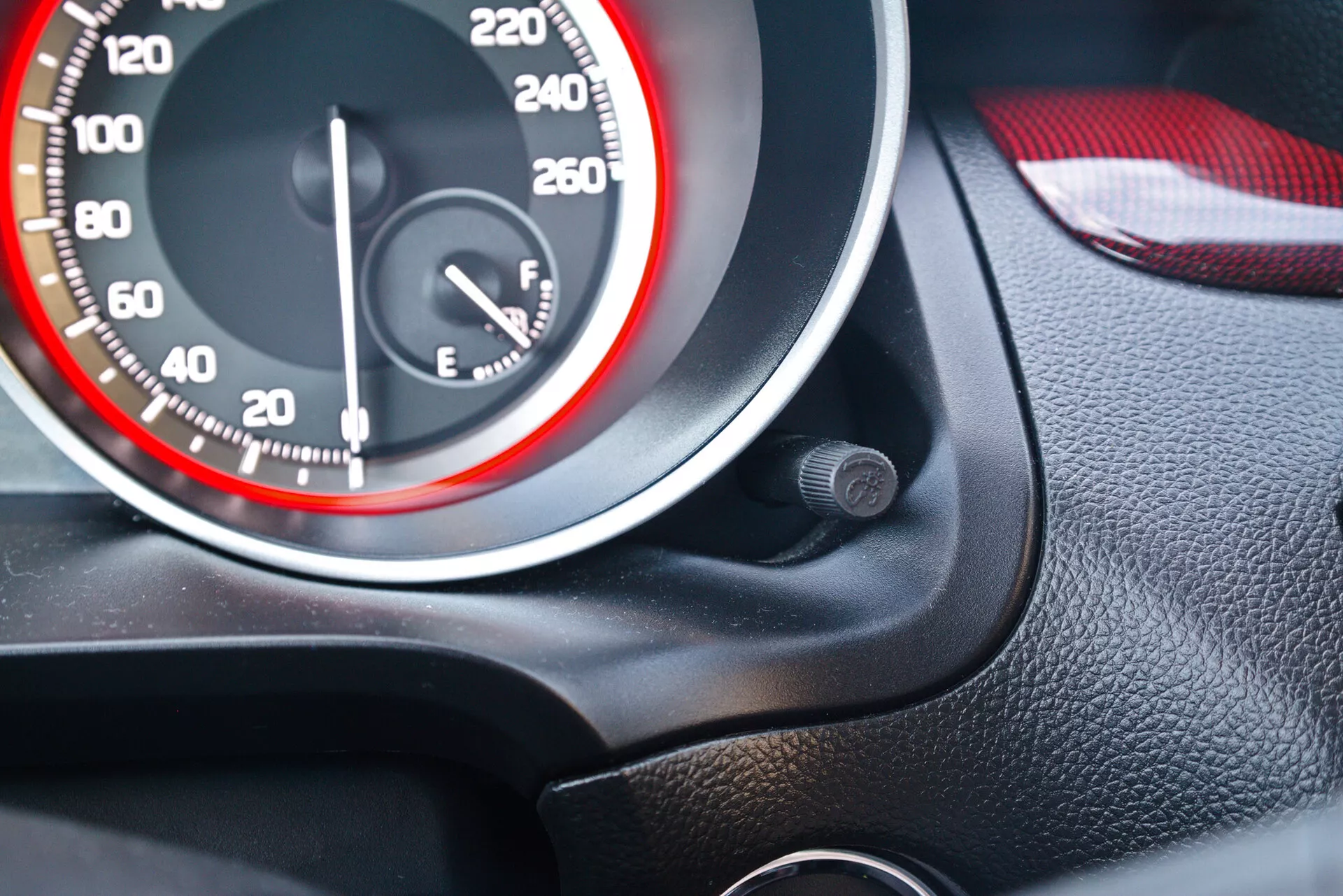 Od blizu prikazuje nadzorno ploščo Suzukija Swift Sport 2017, s poudarkom na merilniku hitrosti in merilniku goriva. Merilnik hitrosti ima bele številke v korakih po 20, doseže do 260 km/h ali mph, kazalec pa je na ničli. Dizajn ima športno rdeče-belo temo z rdečim krogom na notranjem obodu številčnice. Merilnik goriva, ki je skoraj poln, je na desni strani. Posnetek je pod kotom s sedeža voznika s črno zrnavo teksturo okoli merilnikov in barvno usklajenim rdeče-črnim notranjim delom vrat, verjetno iz usnja in plastike. Blizu merilnika hitrosti je majhen okrogel objekt.