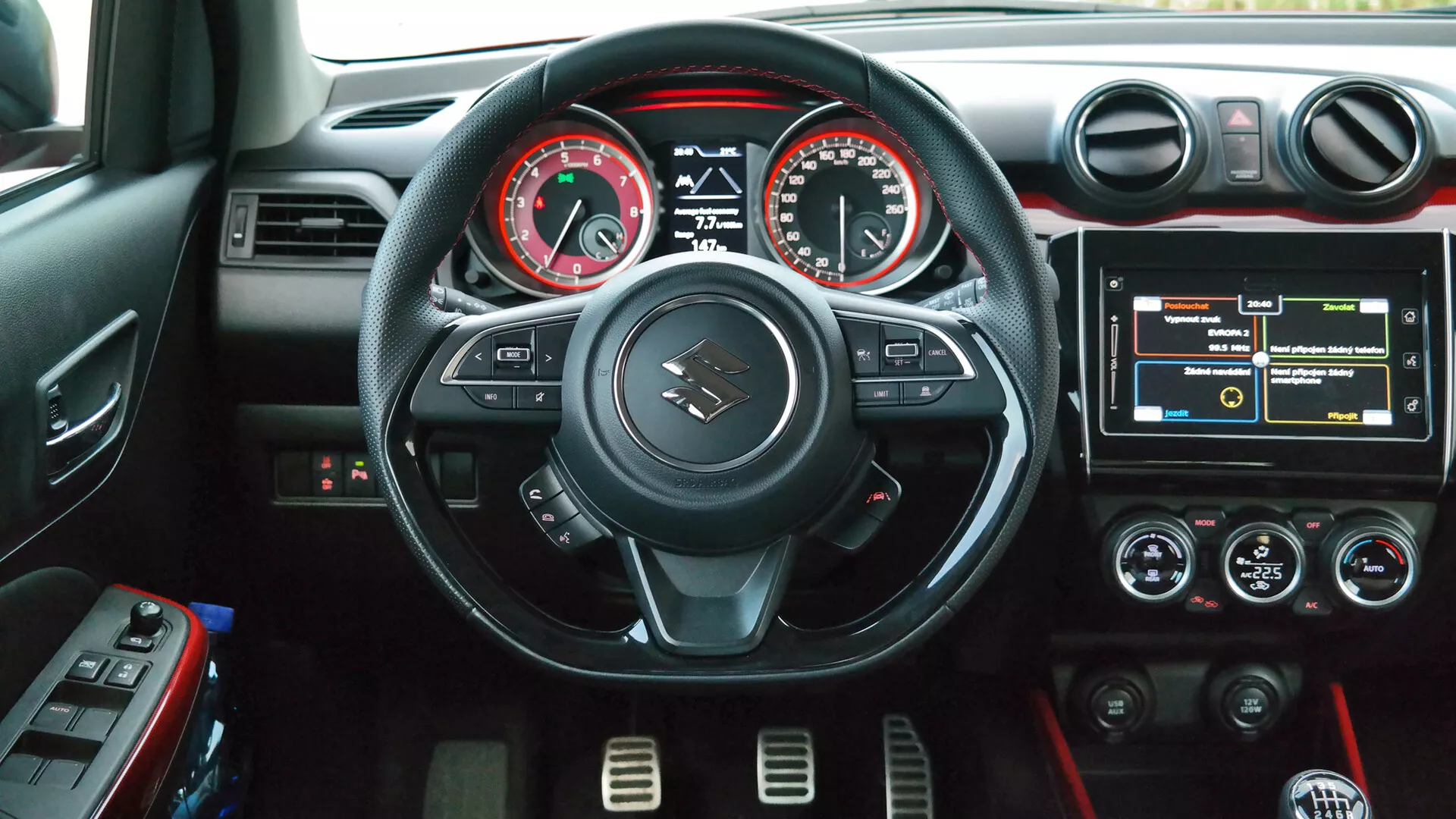 Notranji posnetek Suzuki Swift Sport 2017 iz voznikove perspektive, s trikraki volanom z ravnim dnom s rdečimi šivi in gumbi za upravljanje, analognim tahometrom in merilnikom hitrosti s centralnim digitalnim zaslonom, zaslonom na dotik za informacijski sistem, dvopodročnim nadzorom klimatskih naprav z rdečo ambientalno osvetlitvijo, okroglimi zračniki s srebrnimi poudarki ter držaloma za pijačo na sredinski konzoli.