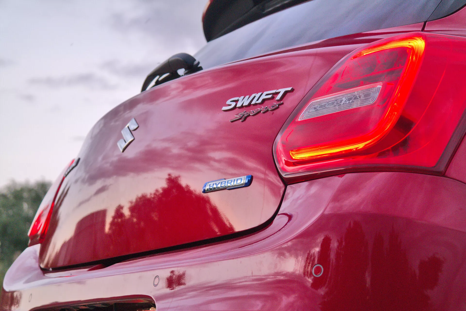 Ta bližinski posnetek prikazuje zadnji desni kot rdečega Suzuki Swift Sport 2017, s poudarkom na značkah vozila in osvetljenem zadnjem svetilniku. Iz nizkega zadnjega kota poudarja zadnje konture vozila, spojler in športno estetiko. Vidni elementi vključujejo emblem Suzuki 'S', oznaki 'SWIFT' in 'Sport', oznako 'HYBRID', dizajn zadnjega odbijača, lita platišča in odseve, ki nakazujejo sijoč zaključek barve.