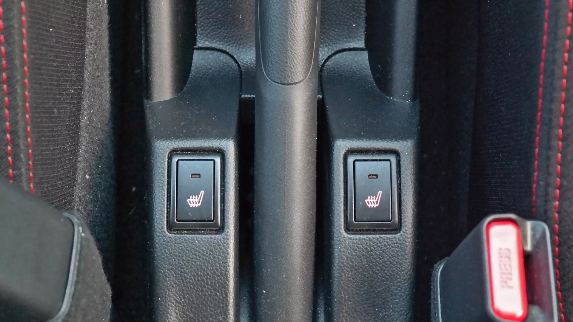 Pogled od blizu na osrednjo konzolo notranjosti Suzuki Swift Sport 2017 z upravljalniki gretja sedežev, teksturirano črno plastiko, rdečimi šivi in delno vidno rdečo zaponko za varnostni pas.