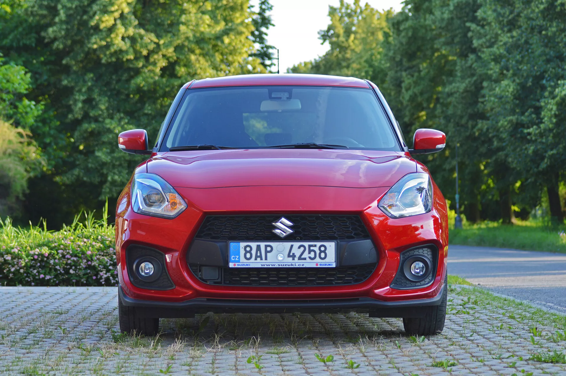 Pogled iz sprednjega kota na rdečo kombilimuzino Suzuki Swift Sport 2017 z izrazito masko in zatemnjenimi žarometi v dnevni naravni svetlobi. Pokrov motorja avtomobila prikazuje aerodinamične linije, meglenke pa povečujejo širino in športnost. Podrobnosti vključujejo možno tehnološko napravo na vetrobranu, večkraka aluminijasta platišča in vidno registrsko tablico. Vozilo je parkirano v primestnem ali parkovnem okolju z drevesi in vegetacijo.