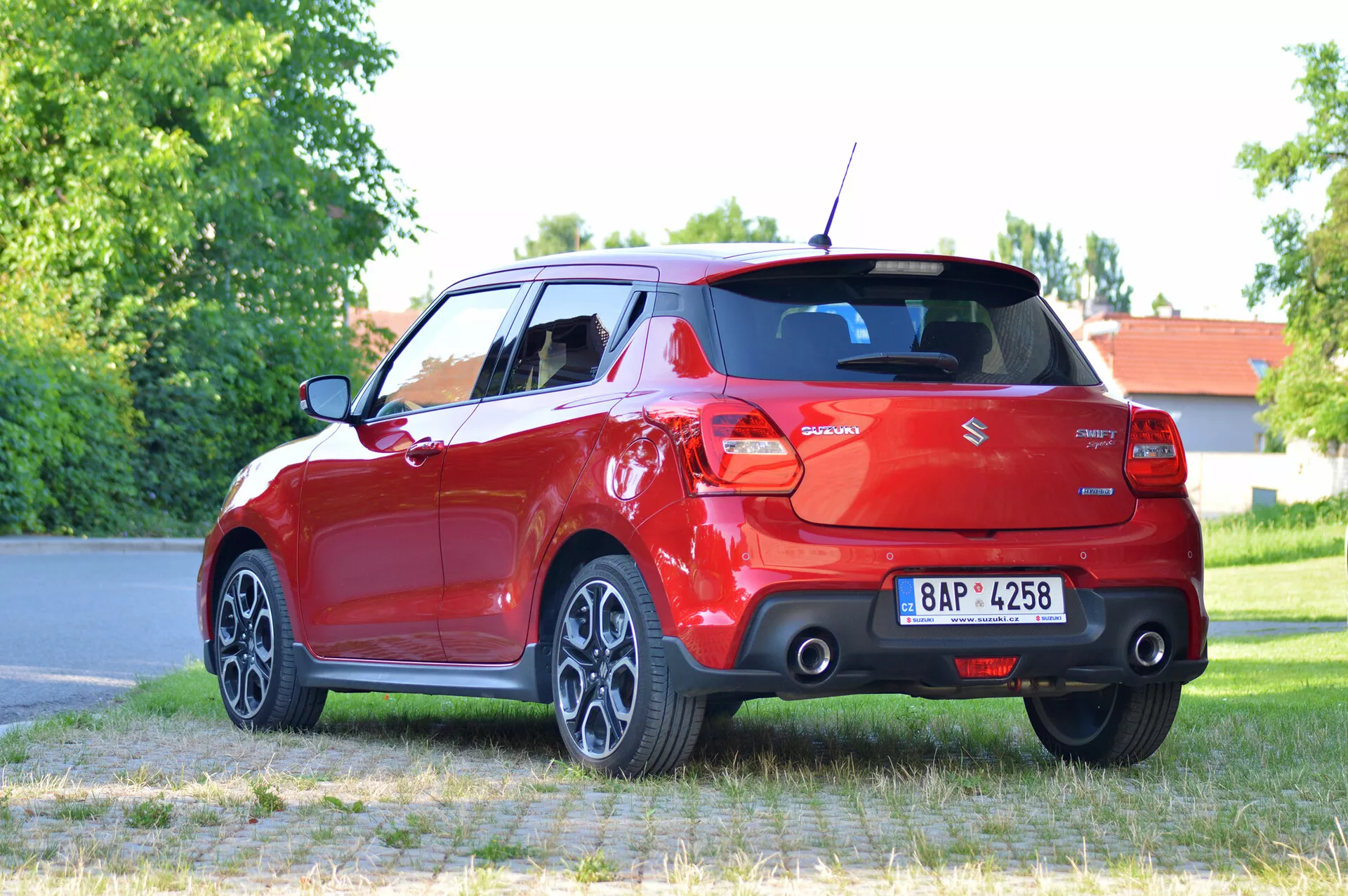 Podrobna slika modernega rdečega hatchbacka Suzuki Swift Sport 2017, nameščenega na tlakovanem površju z travo in drevesi v ozadju. Fotografija je posneta iz tričetrtinskega zadnjega kota, prikazuje desno zadnjo stran avtomobila. Vidne lastnosti vključujejo zadnje luči, zadnje steklo, odbijač, dvojni izpušni sistem, večkraka lita platišča in zavito zadnje kolo. Registrska tablica je zabrisana zaradi zaščite zasebnosti.
