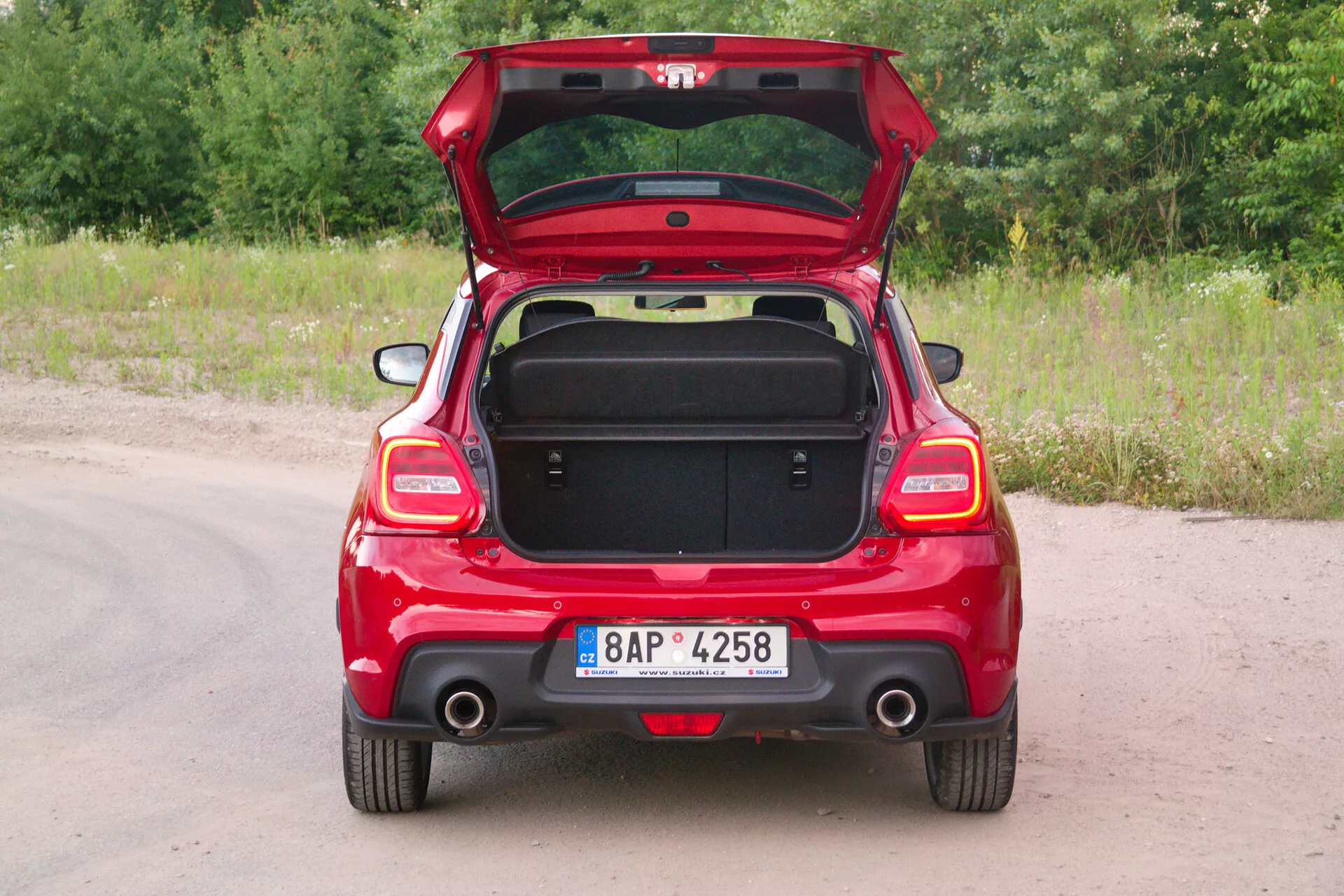 Zadnji pogled na rdeč hatchback Suzuki Swift Sport 2017 z odprtim pokrovom prtljažnika, ki prikazuje simetrične zadnje funkcije, zadnje luči, en sam viden izpušni sistem na strani potnika in zadnji brisalec. Vozilo je parkirano na neasfaltirani poti s travo in zelenjem v ozadju. Registrska tablica je nameščena na sredini zadnjega odbijača. Prazni prtljažnik razkriva skladiščno zmogljivost in notranjo oblogo, z ročajem na notranji strani pokrova za ergonomsko uporabo. Delno zloženi zadnji sedeži nakazujejo delitev 60/40 za razširljiv tovorni prostor. Enostaven zadnji odbijač nakazuje standardne funkcije brez naprednih tehnoloških dodatkov.