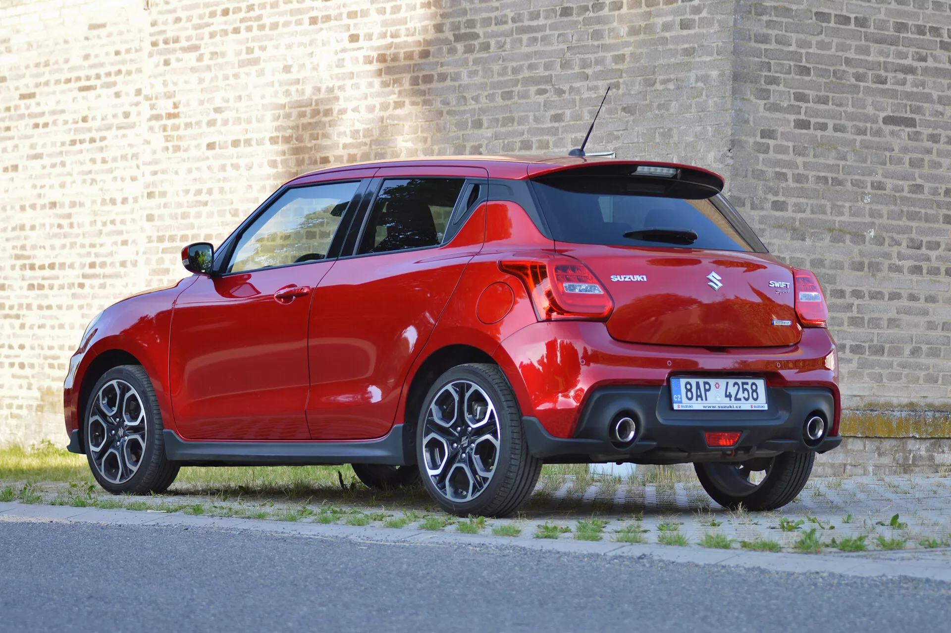 Tričetrtinski zadnji pogled na svetlo rdečega Suzuki Swift Sport 2017, ki poudarja sodoben dizajn z elementi, kot so lita platišča, izrazit zadnji strešni spojler, dvojni izpuhi, kratka radijska antena, parkirni senzorji in vidno označevanje. Avto je parkiran ob pločniku z opečnato steno v ozadju, podnevi.