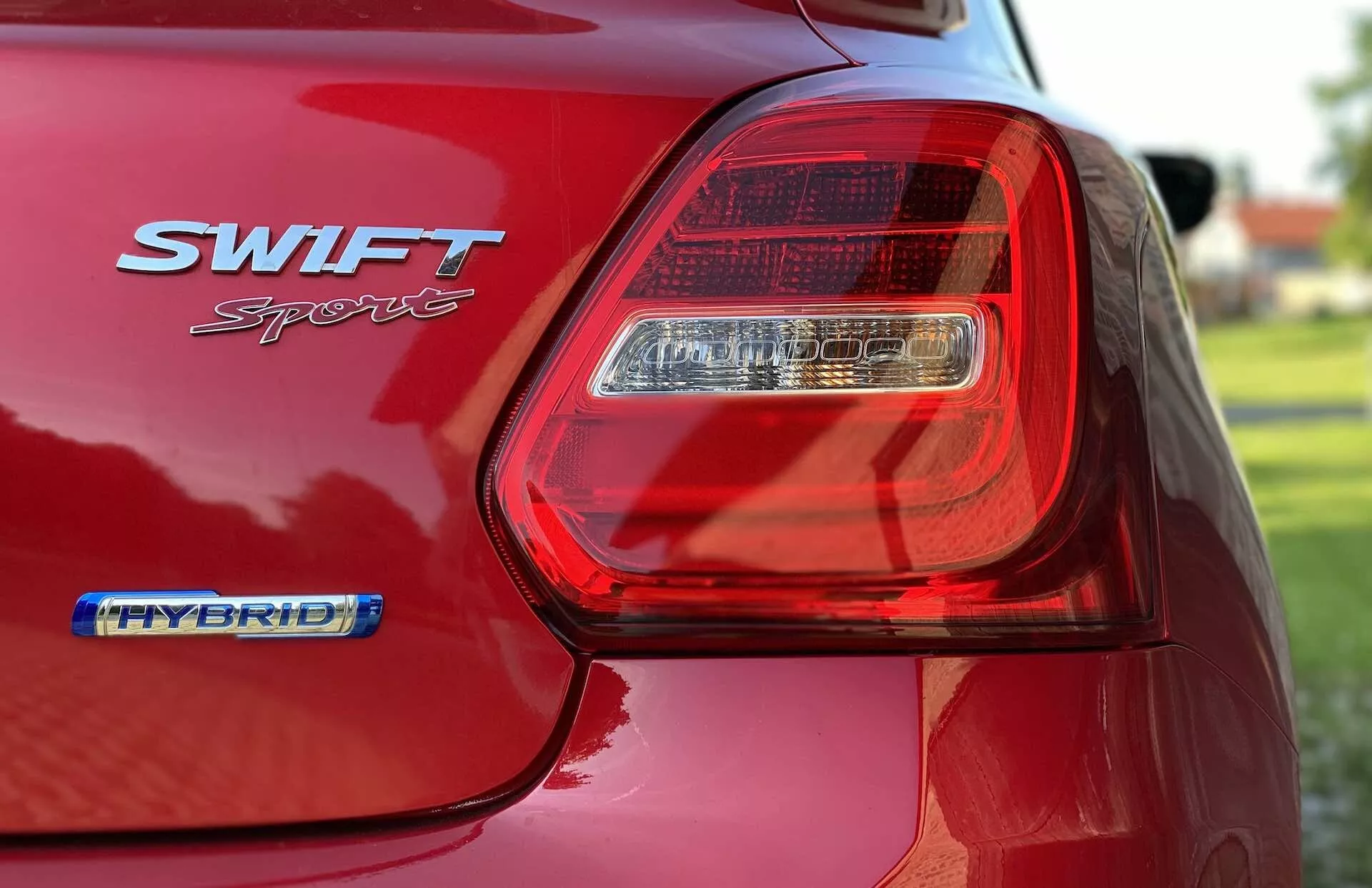 Od blizu pogled na zadnji del rdečega Suzuki Swift Sport 2017, osredotočen na zadnjo LED luč na voznikovi strani. Slika zajema sijoč zaključek barve avtomobila, znački SWIFT Sport in HYBRID ter obrise zadnjega blatnika in dela pokrova prtljažnika. Plitka globinska ostrina zamegli ozadje in poudari podrobnosti svetlobnega sklopa in značk, s poudarkom na moderni zasnovi in hibridni tehnologiji avtomobila.